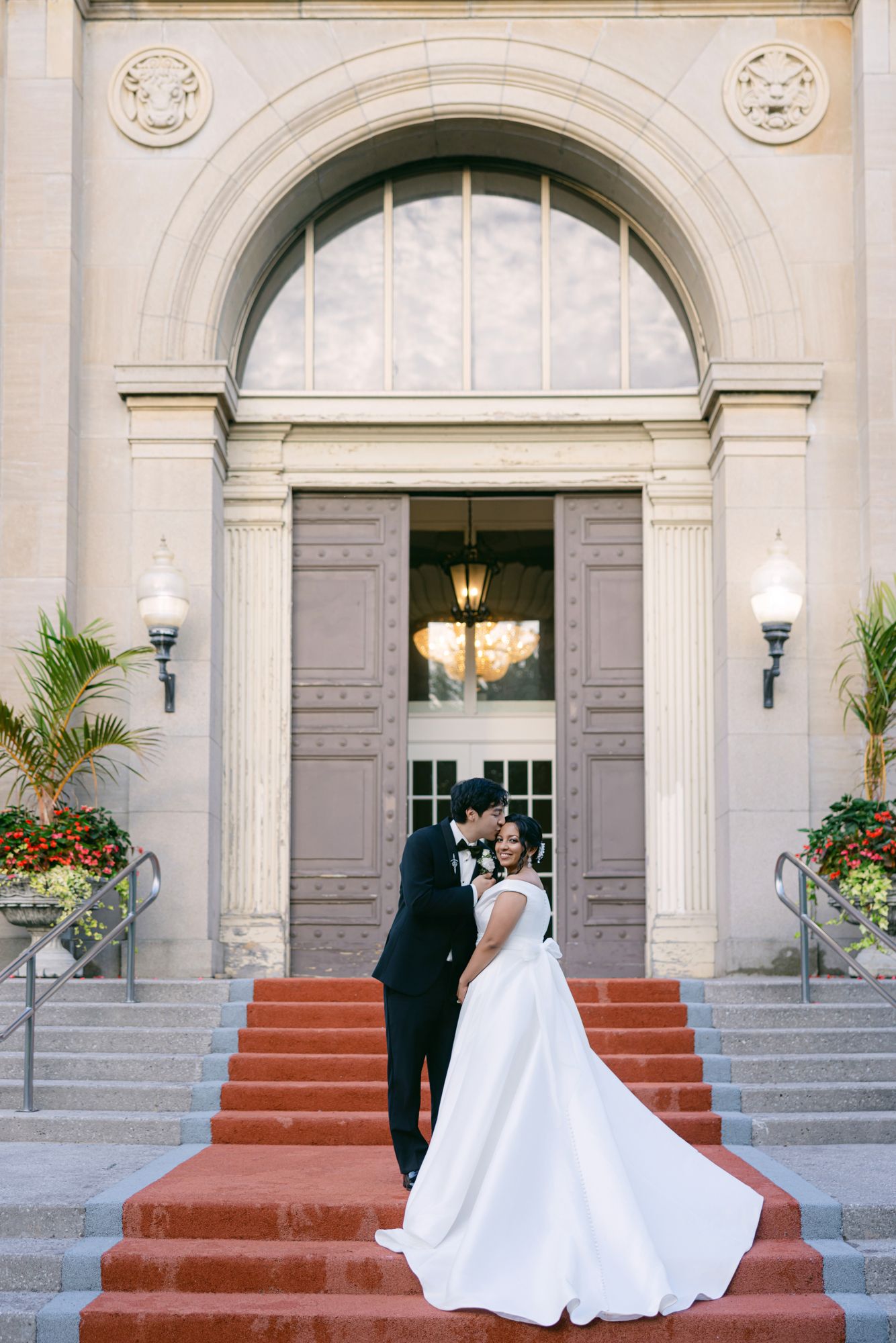 Newlyweds kiss on red-carpeted stone steps outside a grand arched entrance, the bride's long white gown trailing behind.