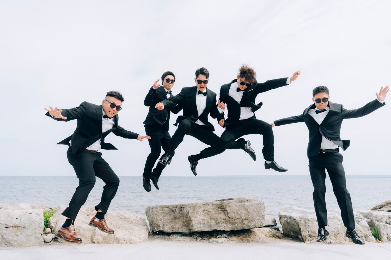 Five groomsmen in black tuxedos jump joyfully on rocks by the sea.