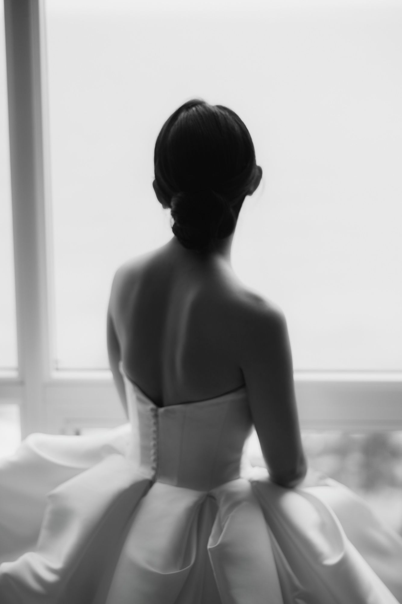 Elegant bride in strapless white ball gown viewed from behind, gazing out window, black and white.