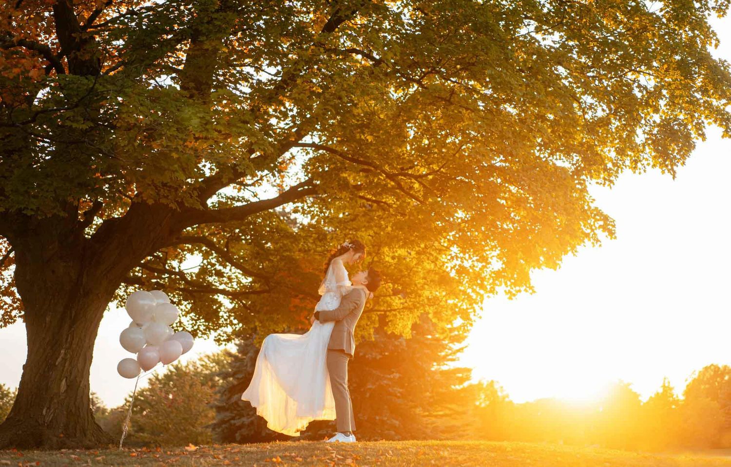 Pregnant woman in white dress holding balloons under golden tree at sunset in a serene outdoor maternity photo.