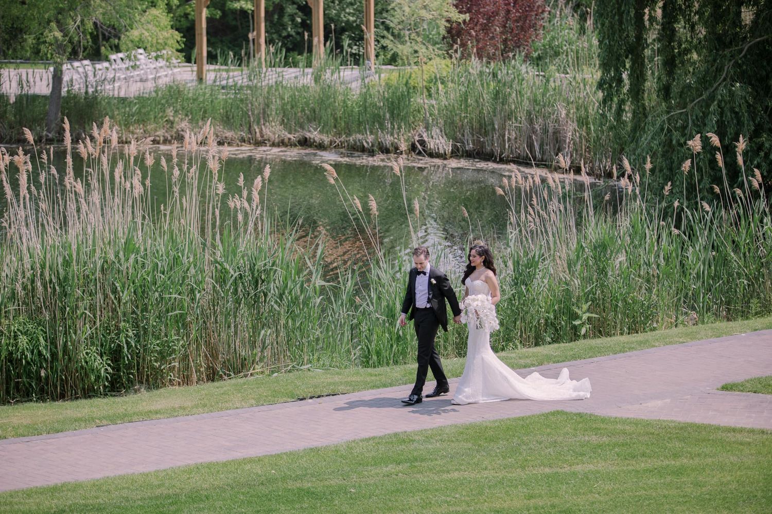 Bride and groom walking hand in hand along scenic garden path beside tranquil pond.