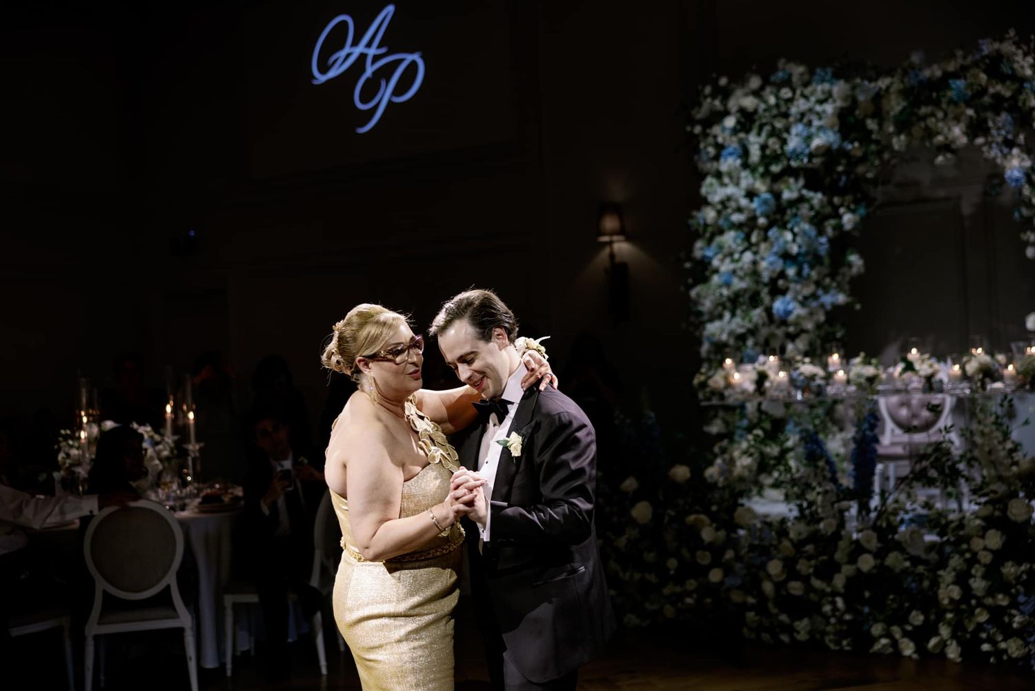 Groom dances with mother in elegant ballroom with monogram lighting at wedding reception.