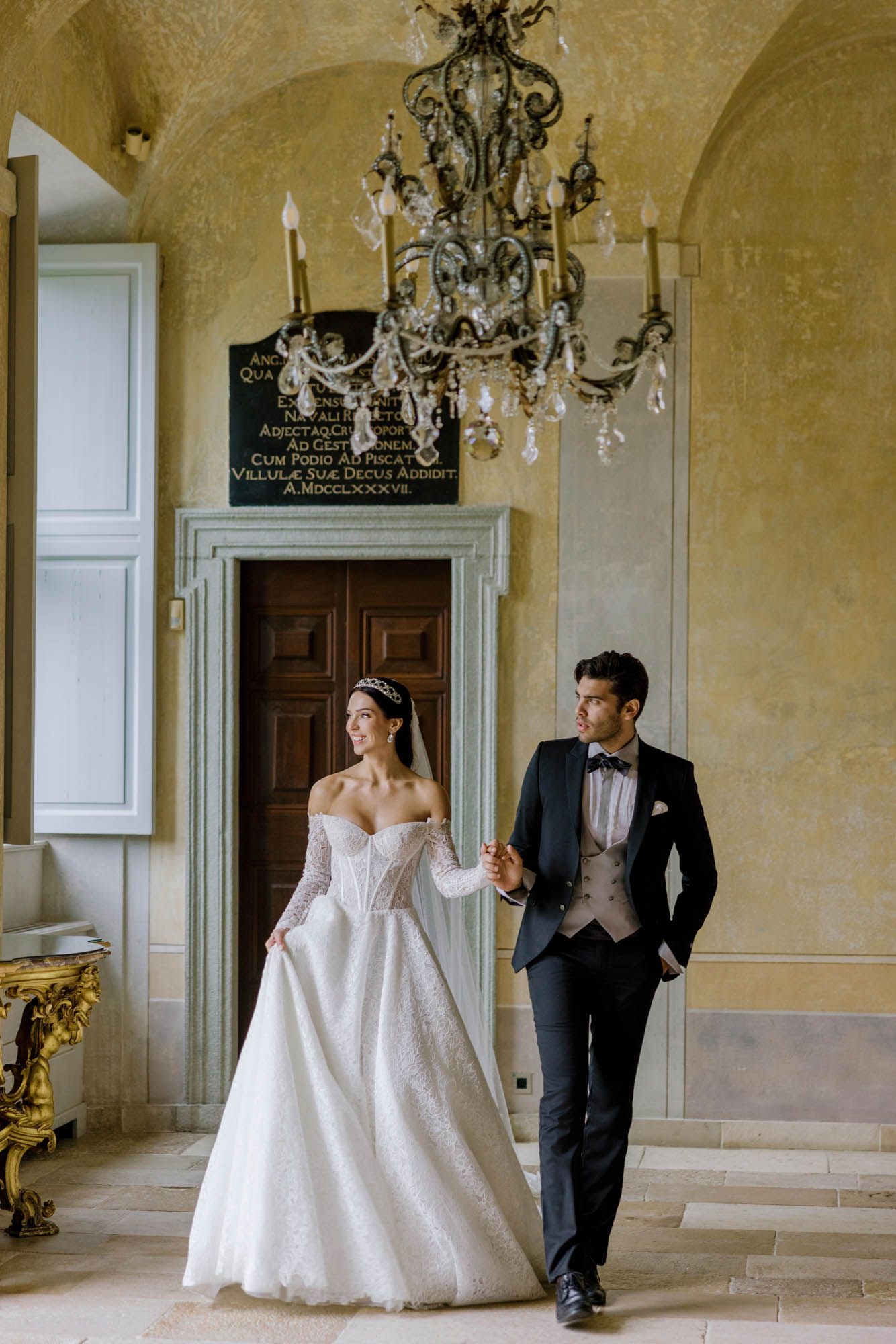Bride and groom pose elegantly in a grand European venue with chandelier and arched ceilings.