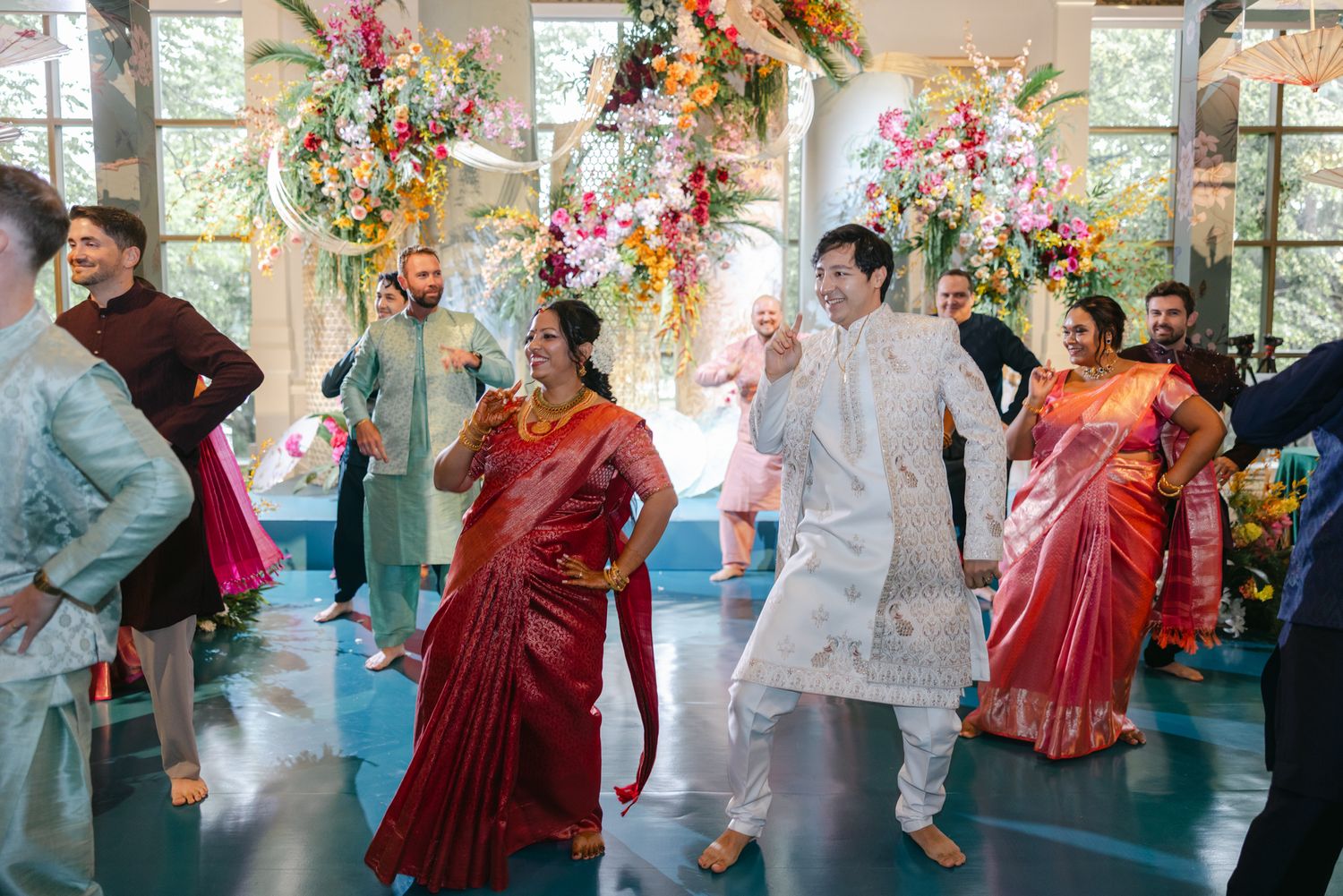 Guests in colorful traditional Indian attire dance joyfully at a wedding celebration beneath lush pink and orange floral arrangements.