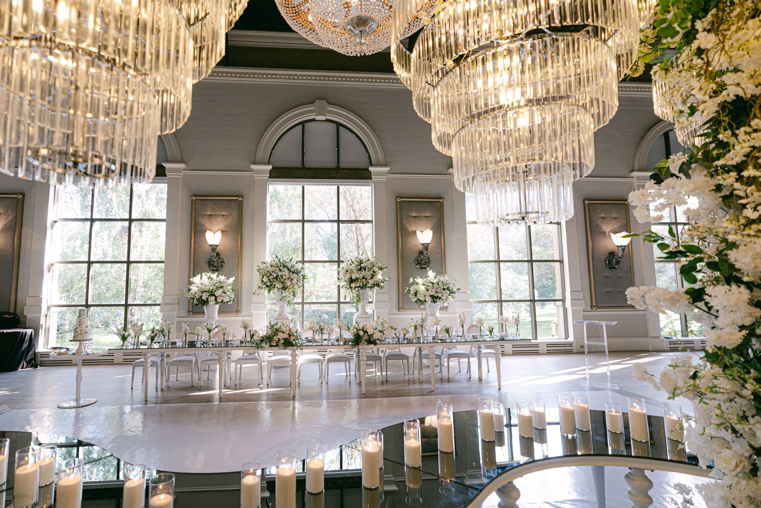 Elegant wedding reception hall with crystal chandeliers, tall arched windows, white floral arrangements and glowing candles.