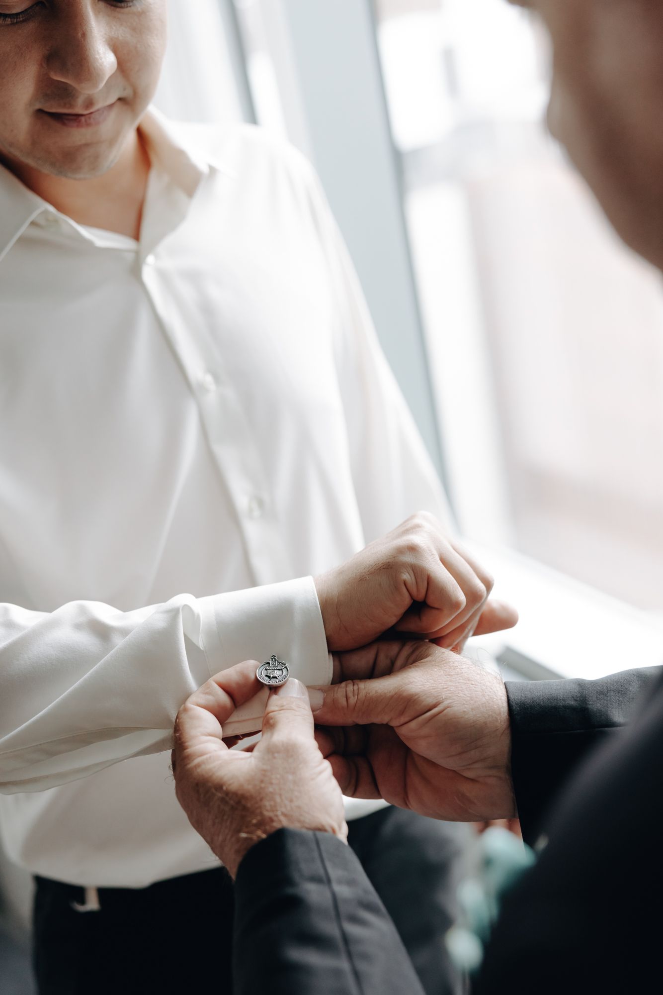 Groomsman in a white dress shirt has a cufflink fastened by another man's hands beside a bright window before a wedding.