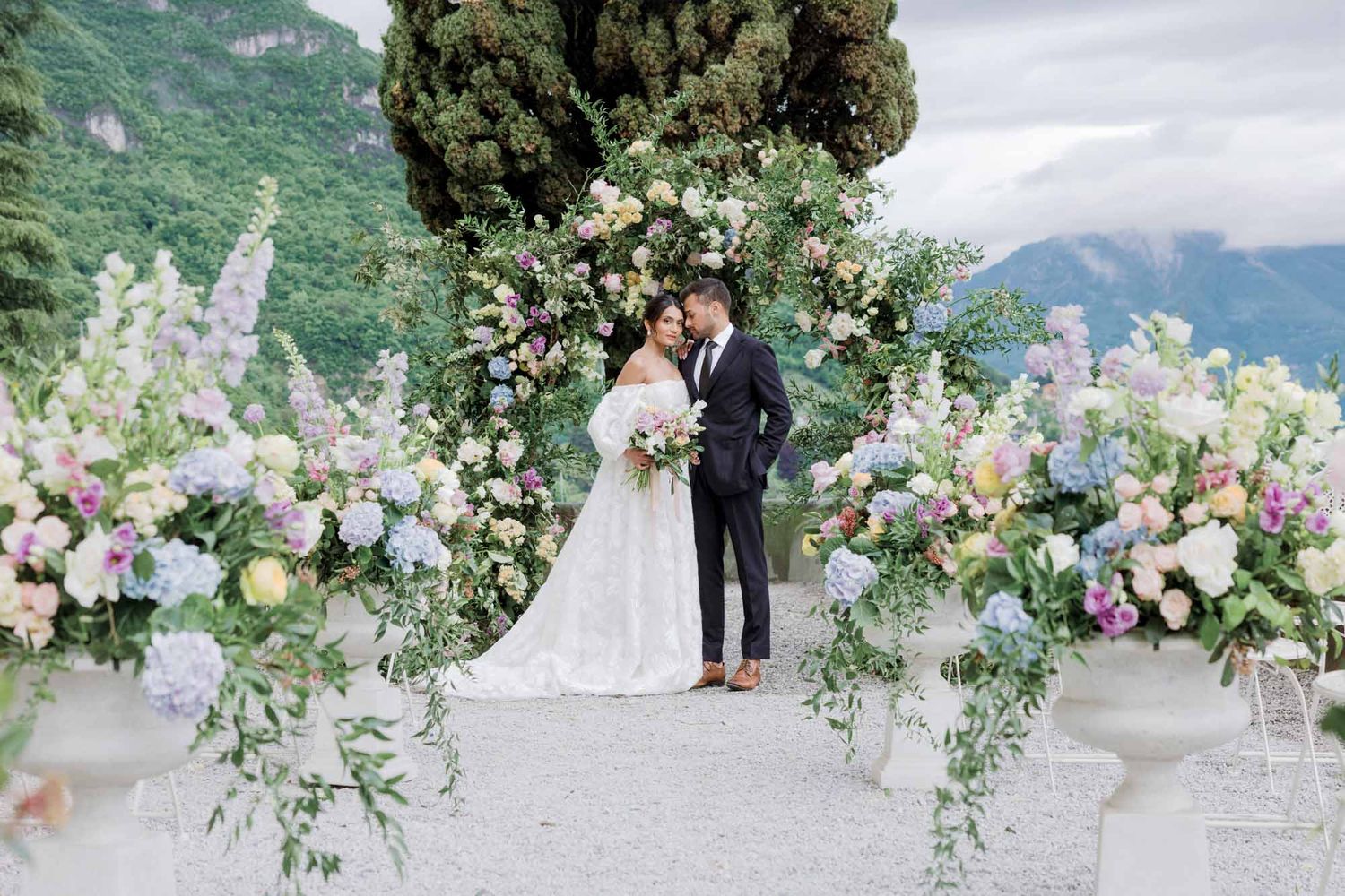 Couple shares a romantic kiss under a lush floral arch with mountain views at Lake Como.