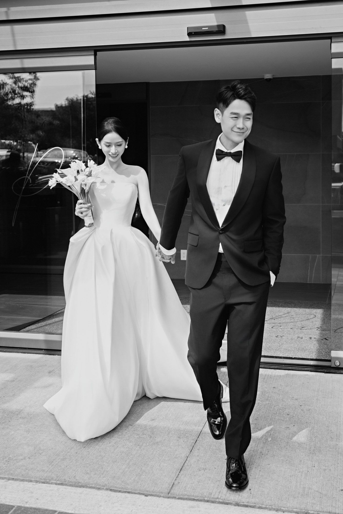 Newlywed couple walks hand-in-hand in black and white photo, bride in ballgown holding lily bouquet.