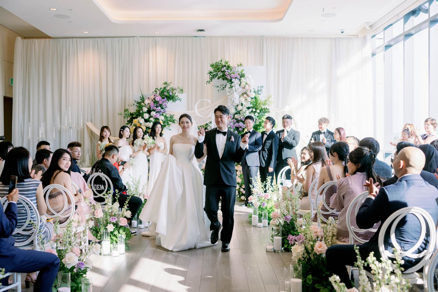 Newlyweds walk hand in hand down aisle past seated guests at elegant floral wedding ceremony.