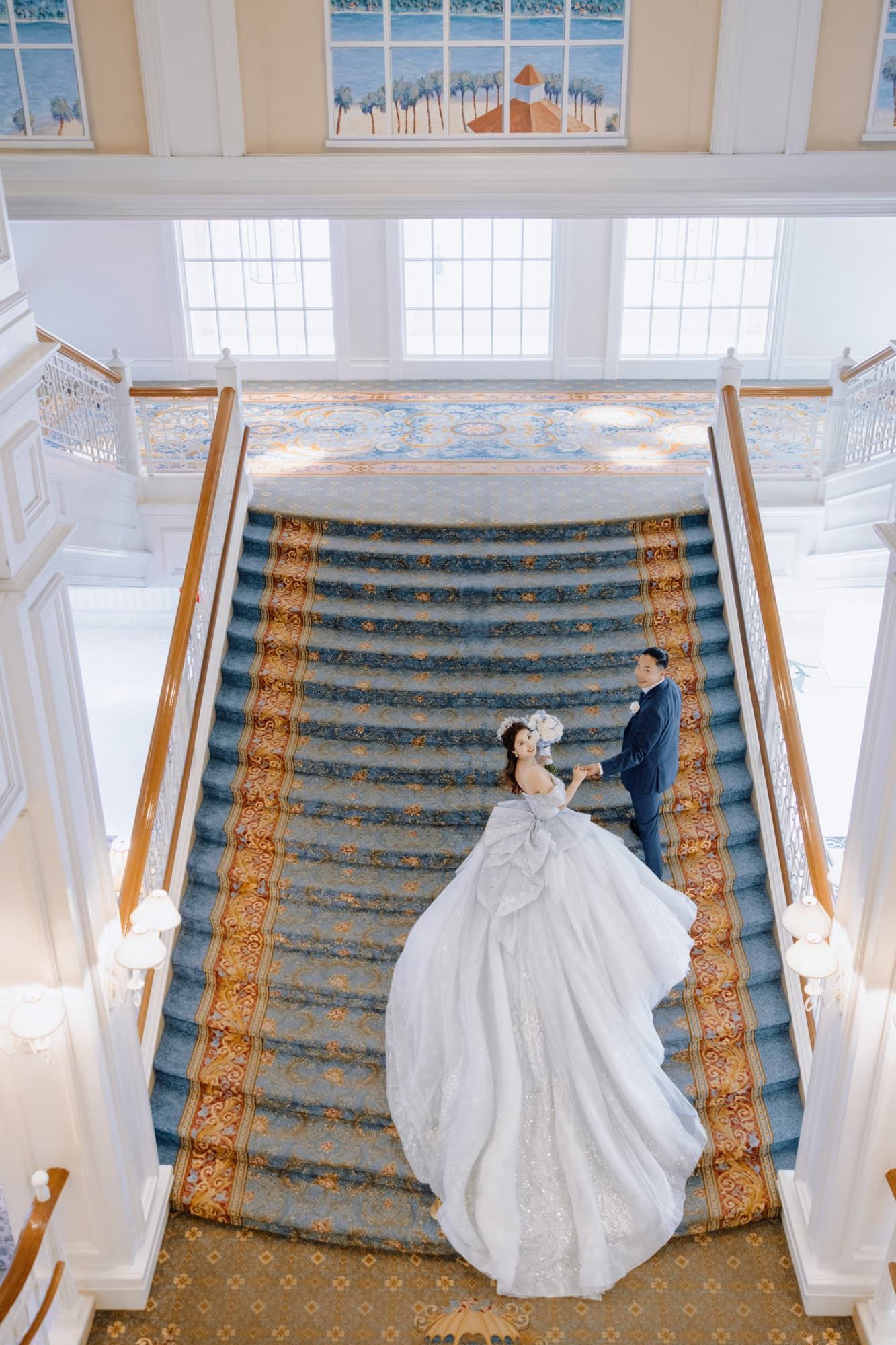 Bride and groom holding hands on elegant grand staircase with blue carpet and ornate gold runner in luxury wedding venue.