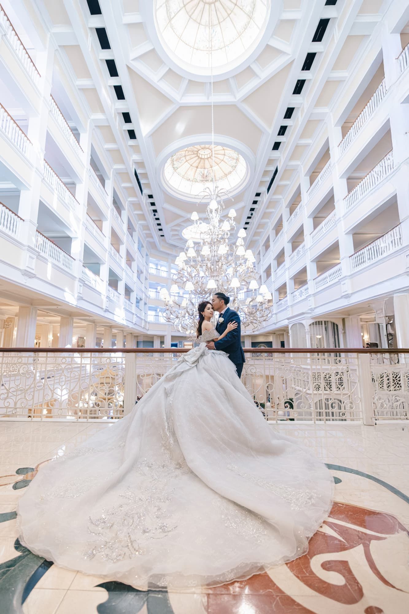 Bride and groom embracing under grand chandelier in elegant white ballroom with sweeping wedding gown.
