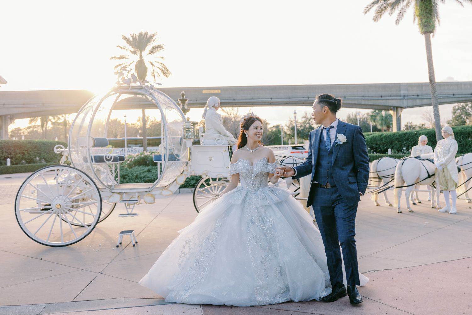 Bride and groom pose together at sunset with a white Cinderella carriage and palm trees in the background.