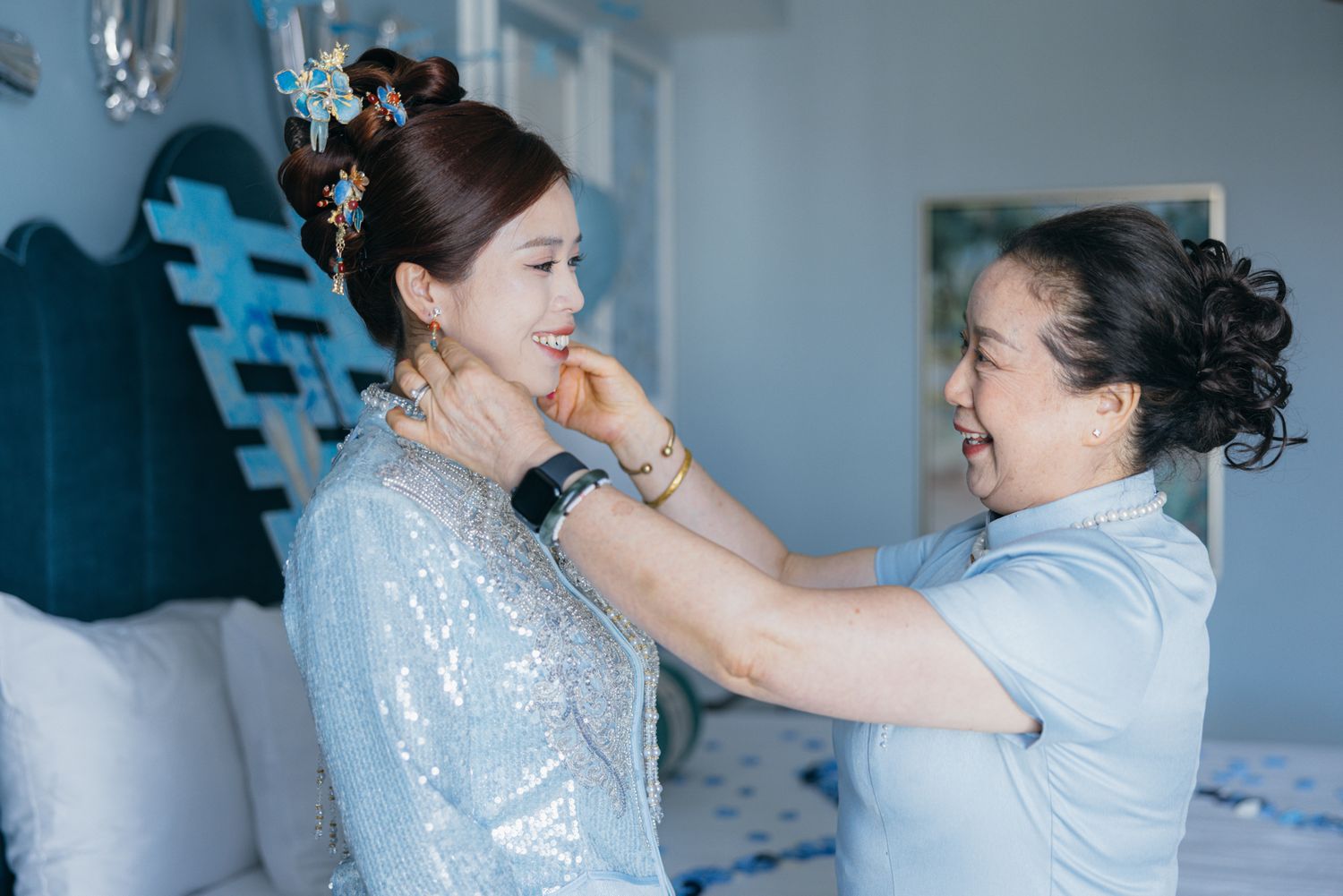 Older woman smiling while fastening a necklace on a bride in a sequined blue dress before her wedding.