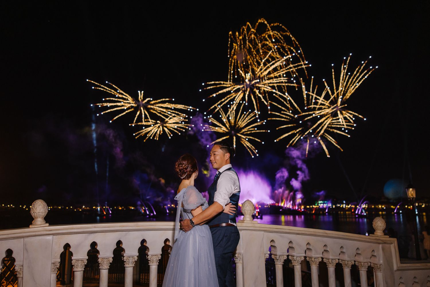 Wedding couple embraces on balcony with spectacular fireworks display lighting the night sky behind them.