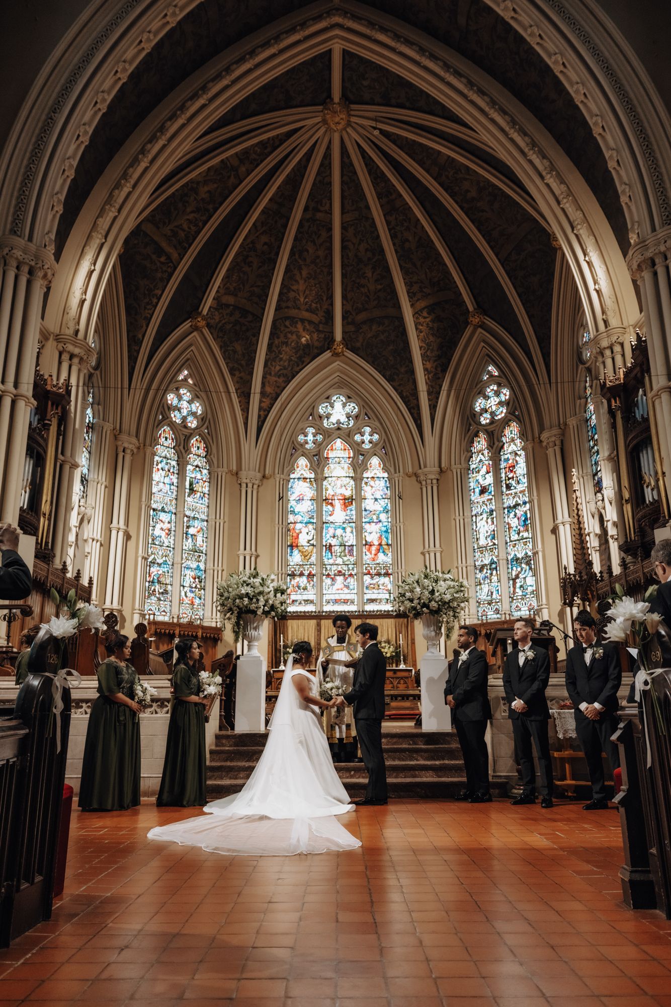 Bride and groom stand at the altar of a Gothic church with stained glass windows, bridesmaids and groomsmen beside them.