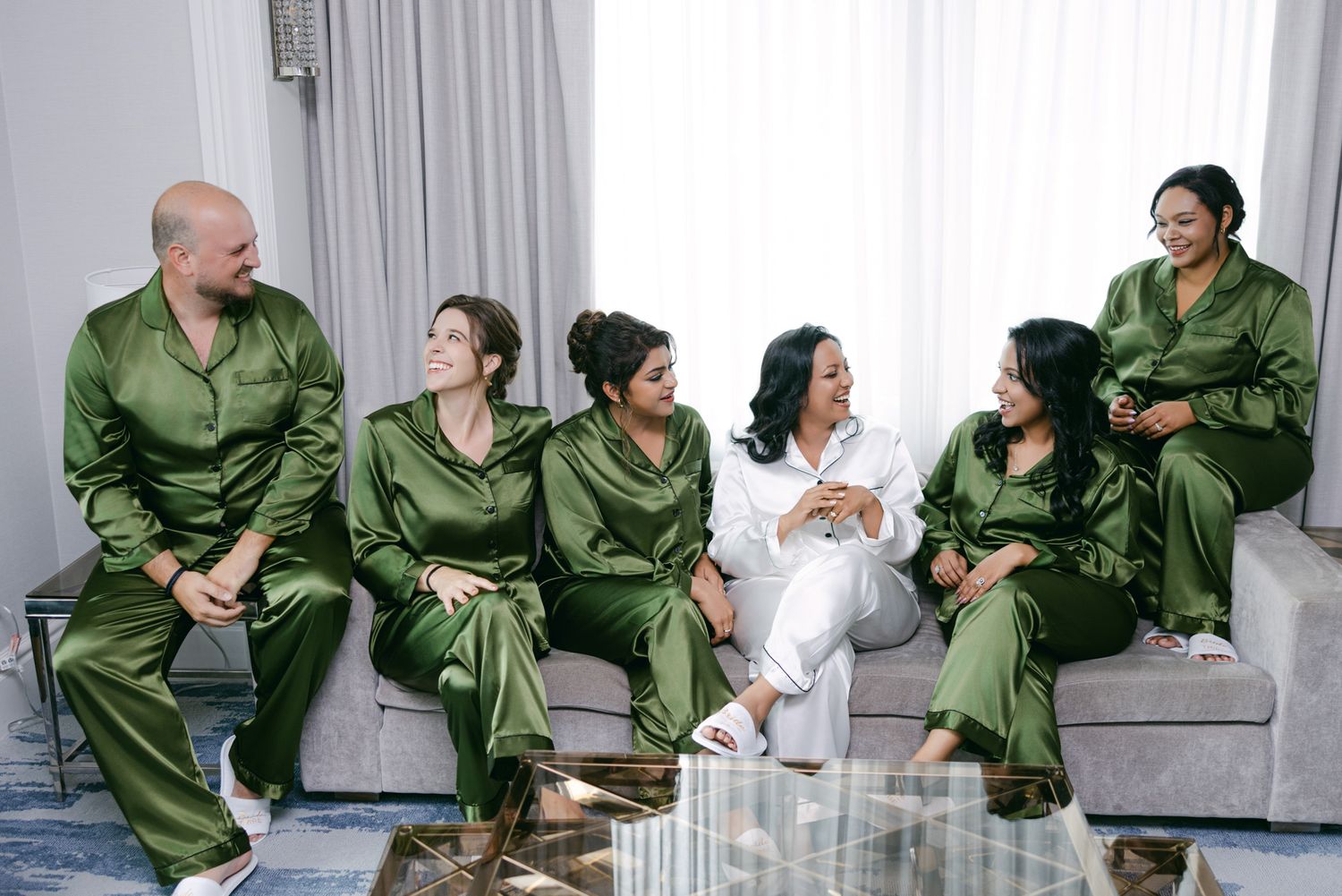 Six people relax on a gray sofa wearing matching olive green satin pajama sets, with one in white at the center, in a bright room.
