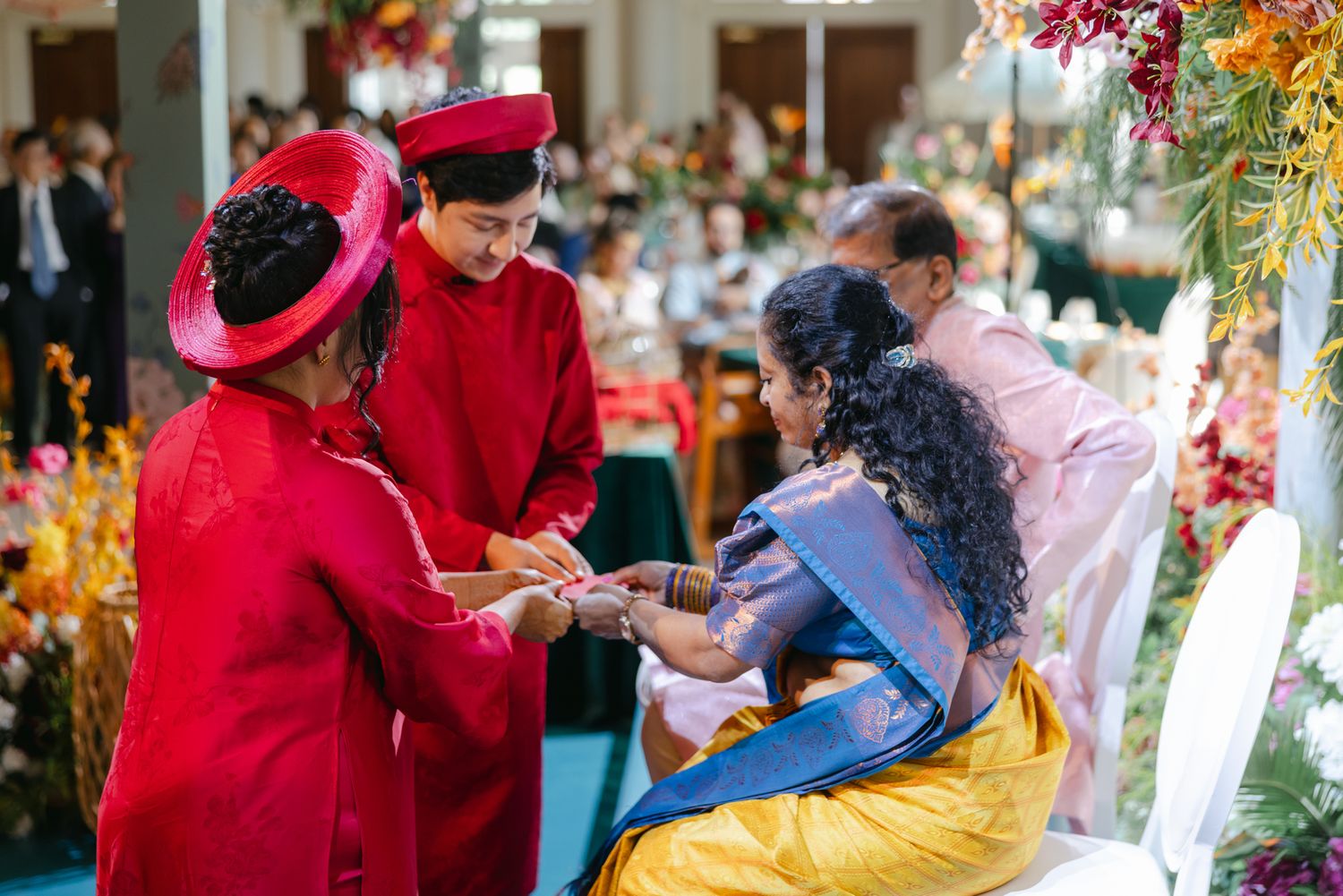 Two people in red traditional ao dai and headwear perform a tea ceremony with a seated woman in a blue sari at a flower-decorated wedding.