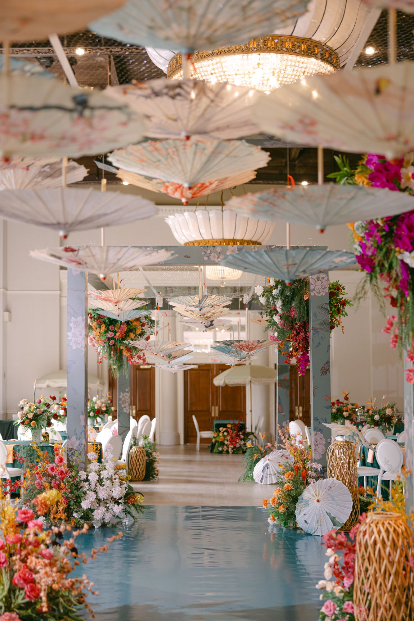 Wedding reception hall with rows of hanging paper parasols, a crystal chandelier, lush floral displays and a reflective blue aisle.