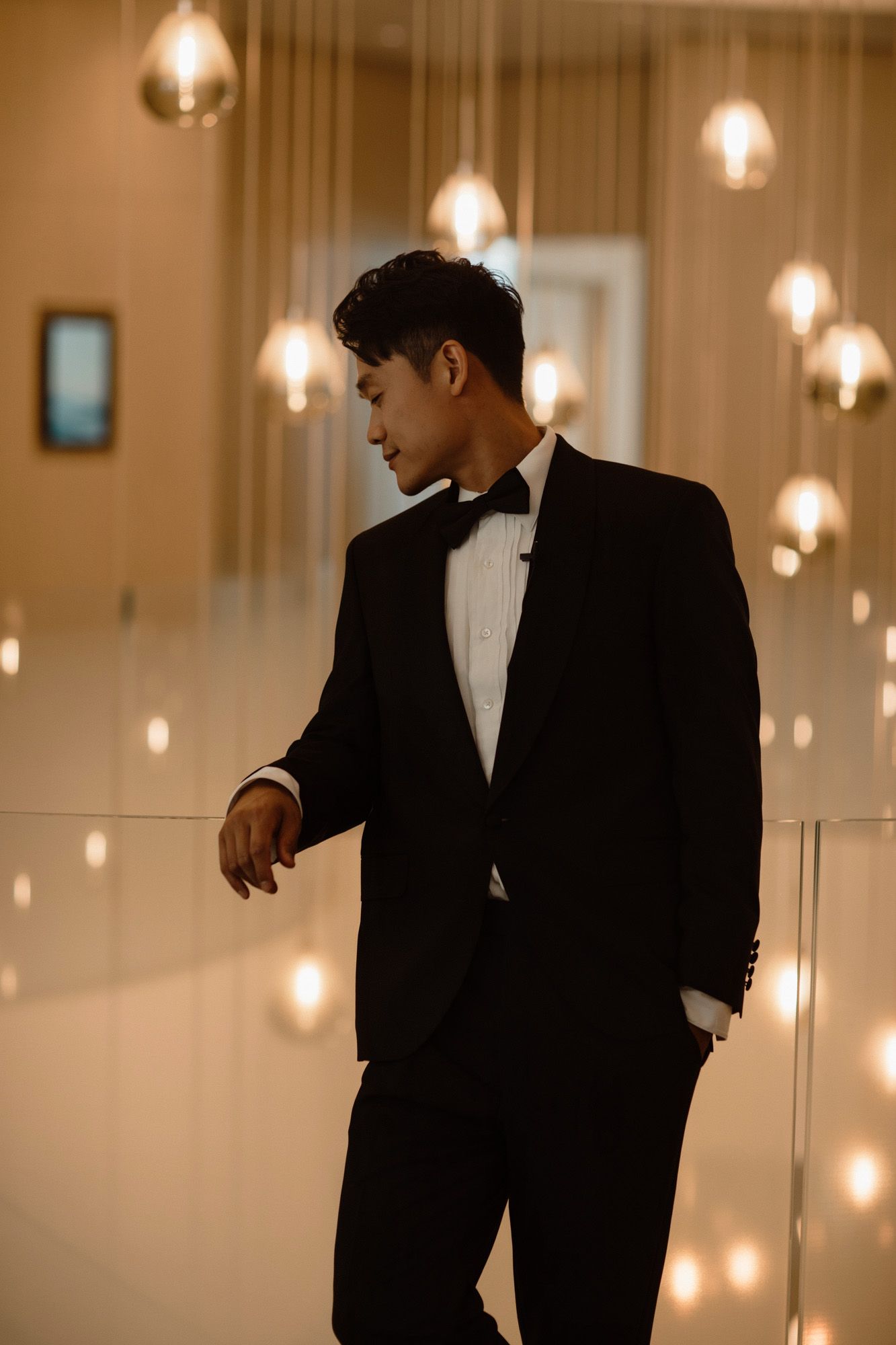 Elegant groom in black tuxedo and bow tie posing under warm pendant lights.