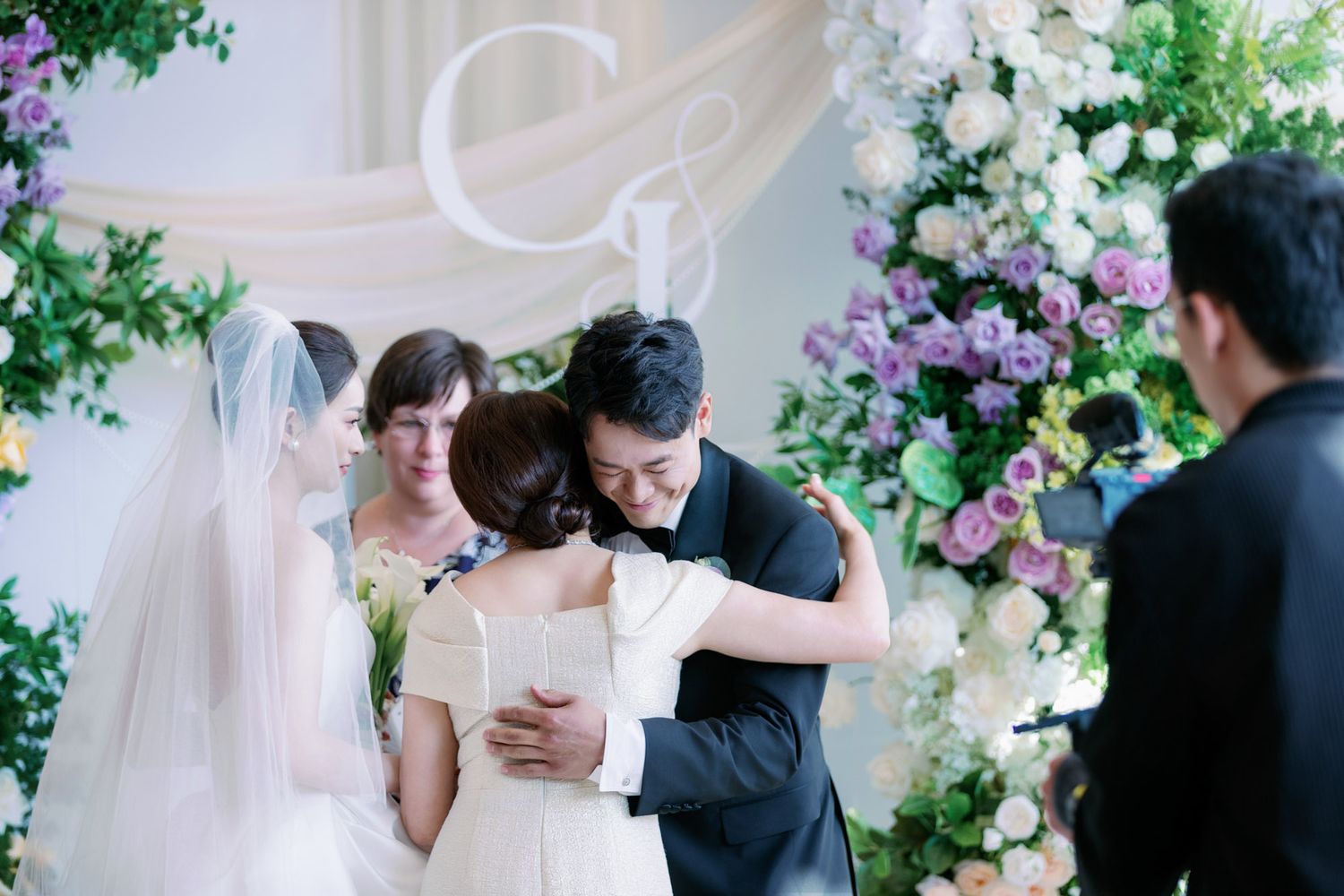 Groom embraces bride and mother in emotional moment at beautifully decorated wedding altar.