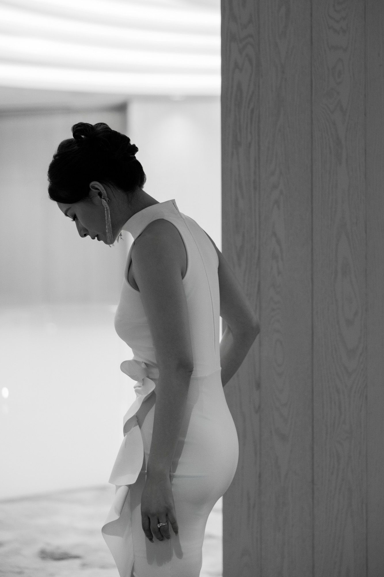 Black and white portrait of bride in fitted white dress with ruffled detail leaning against wall.
