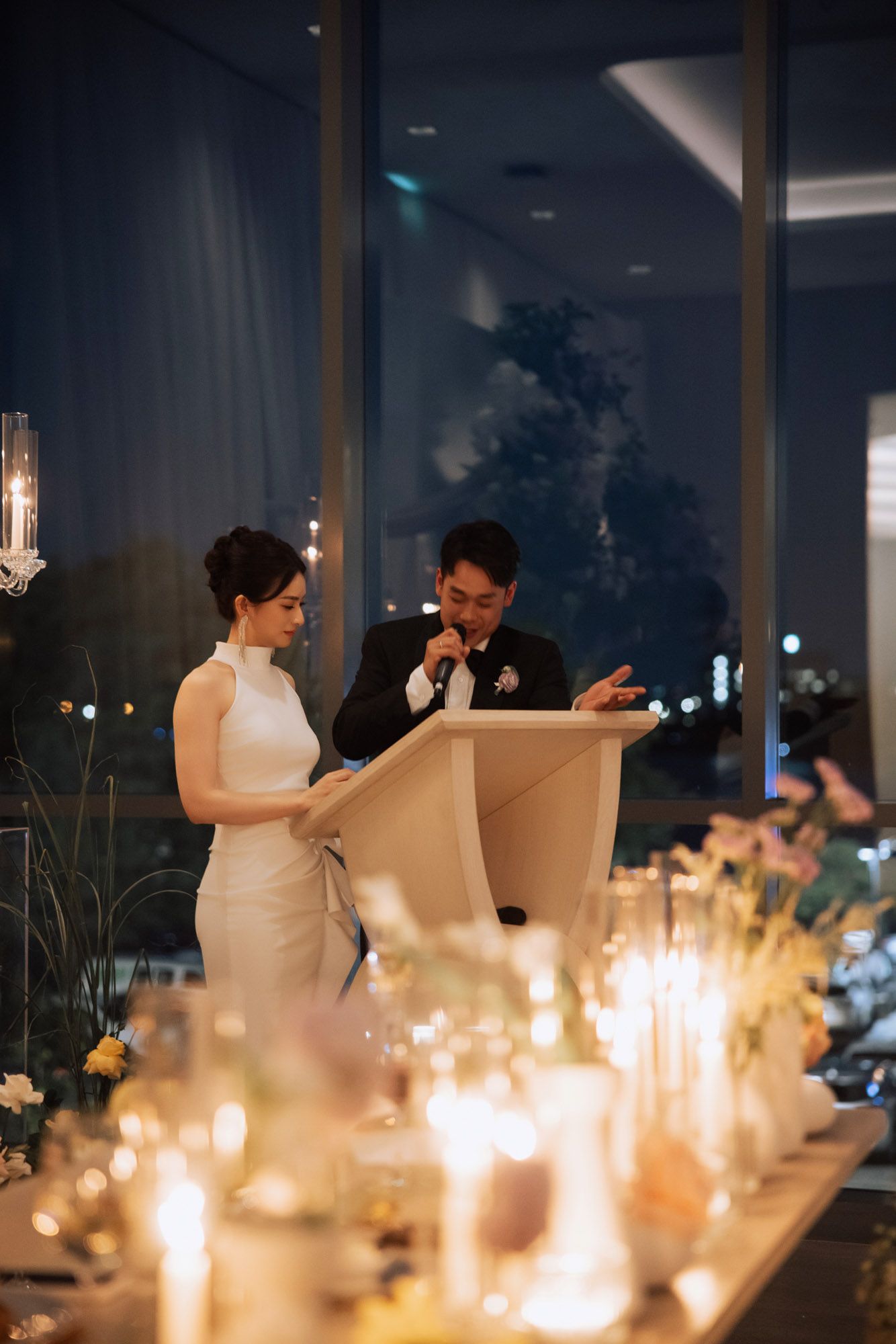 Bride in white gown and groom in tuxedo deliver wedding speeches at a candlelit evening reception.