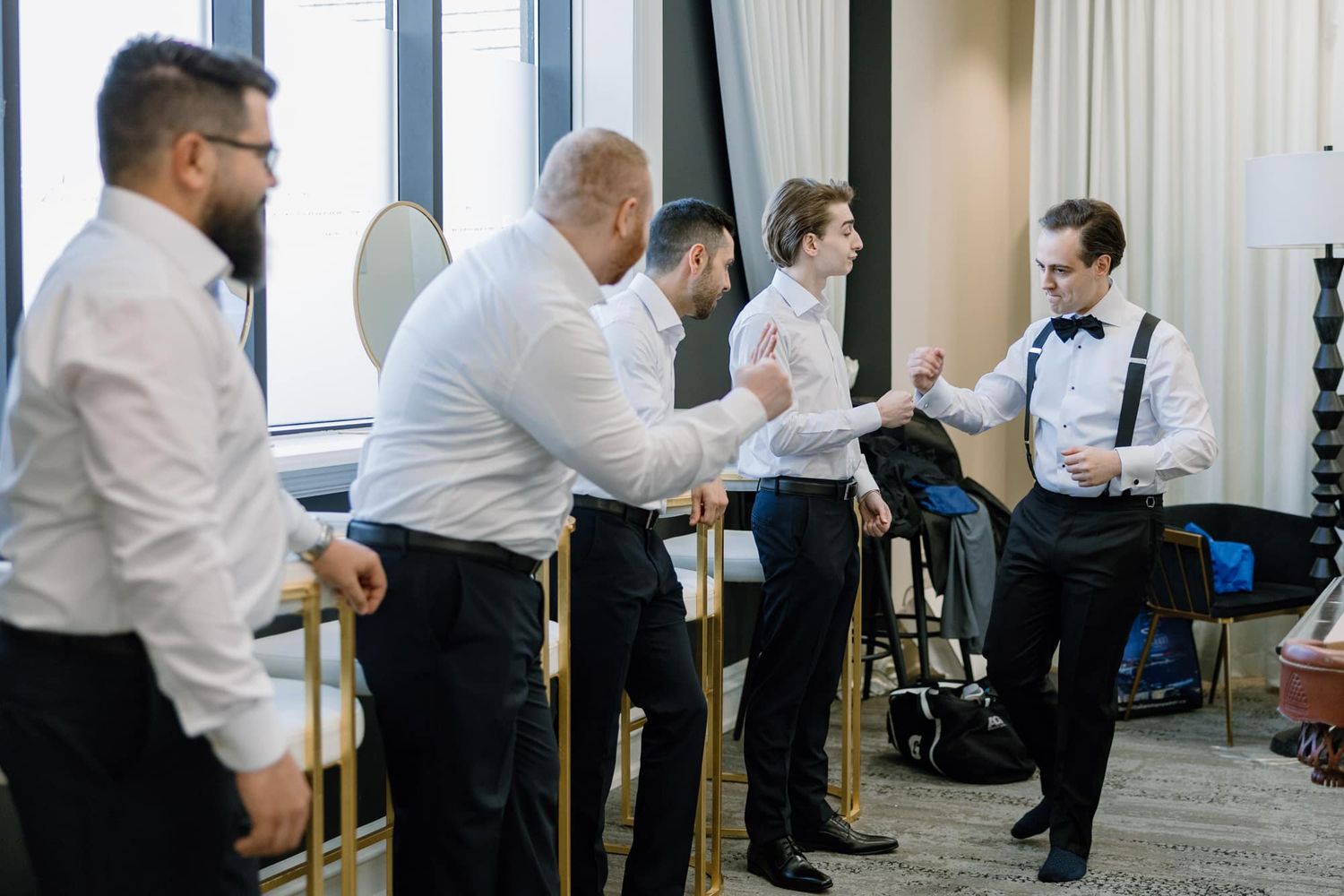 Groomsmen in white shirts and black pants helping each other get dressed before a wedding ceremony.