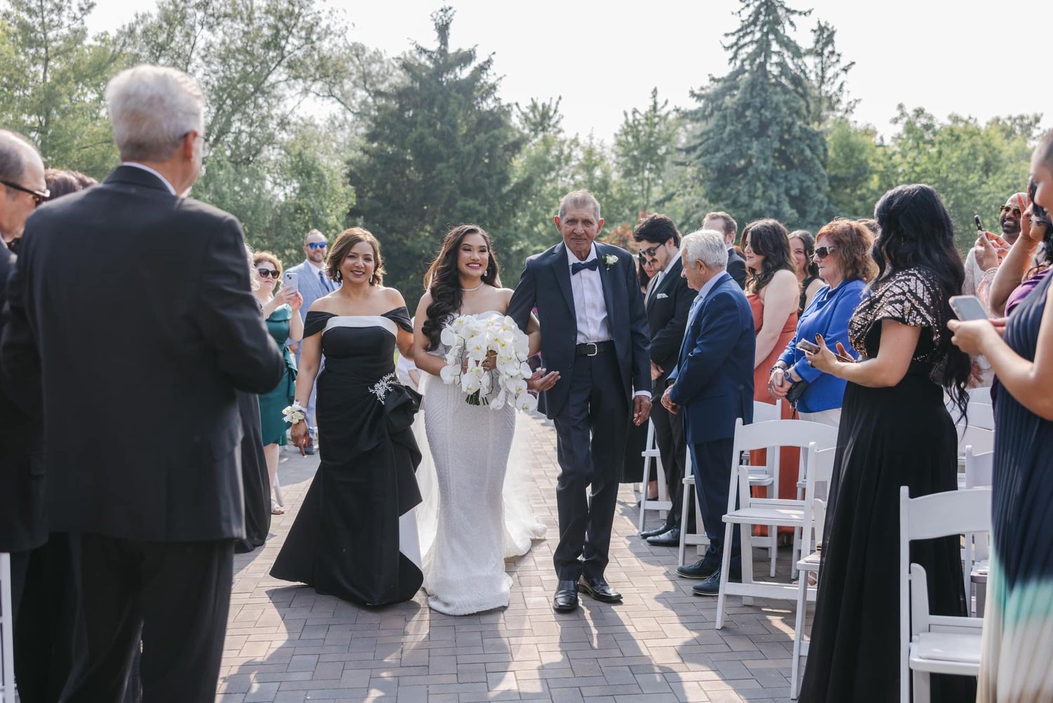 Bride walks down outdoor aisle escorted by parents at sunny garden wedding ceremony.