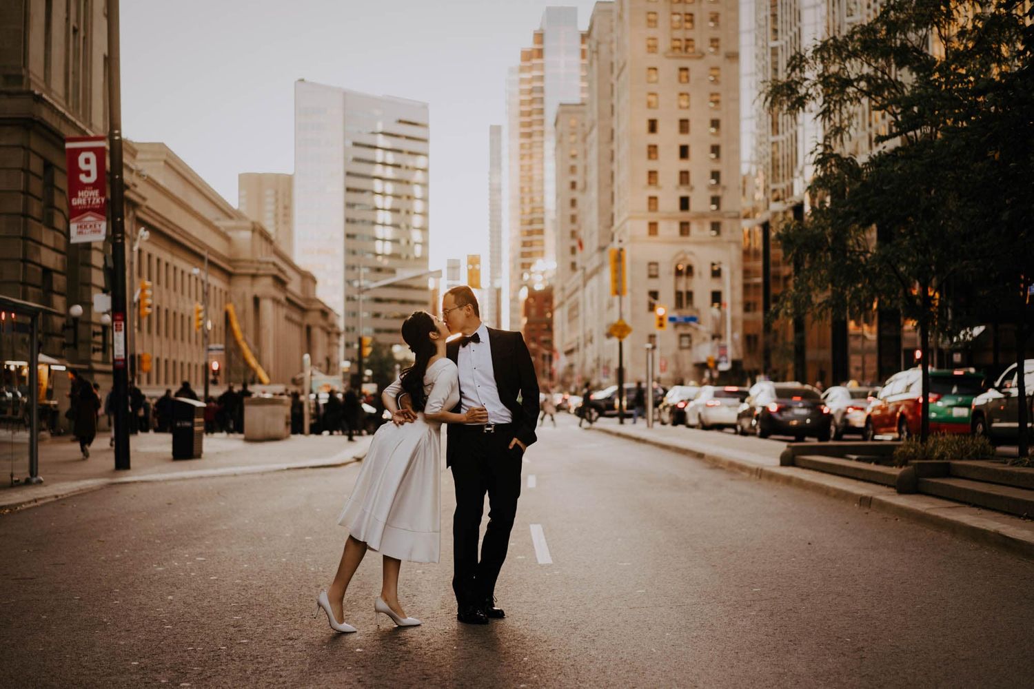 Bride and groom share a romantic kiss in the middle of a golden-lit city street at sunset.