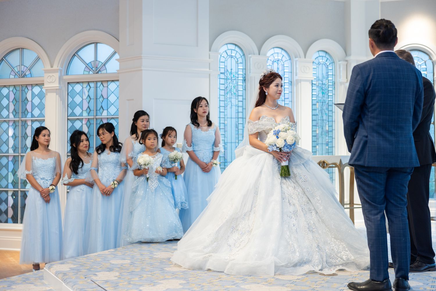 Bride in white ball gown stands at altar with bridesmaids in blue dresses inside elegant chapel.