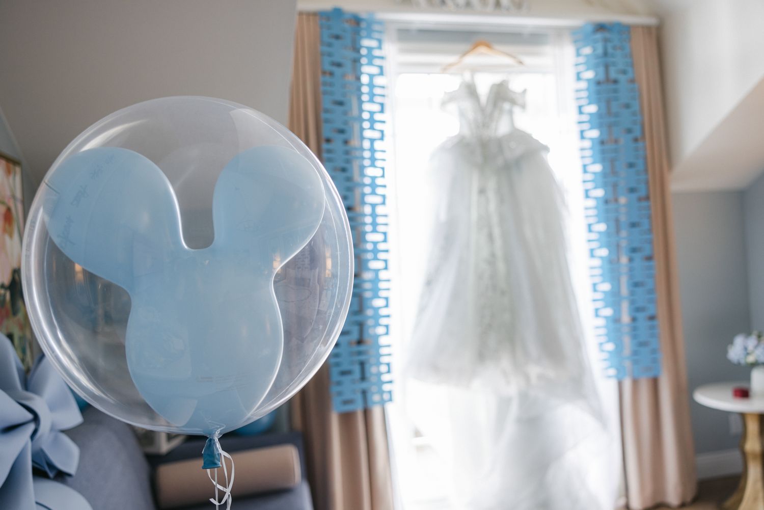A light blue Mickey Mouse balloon floats in front of a wedding dress hanging near a window with blue curtains.