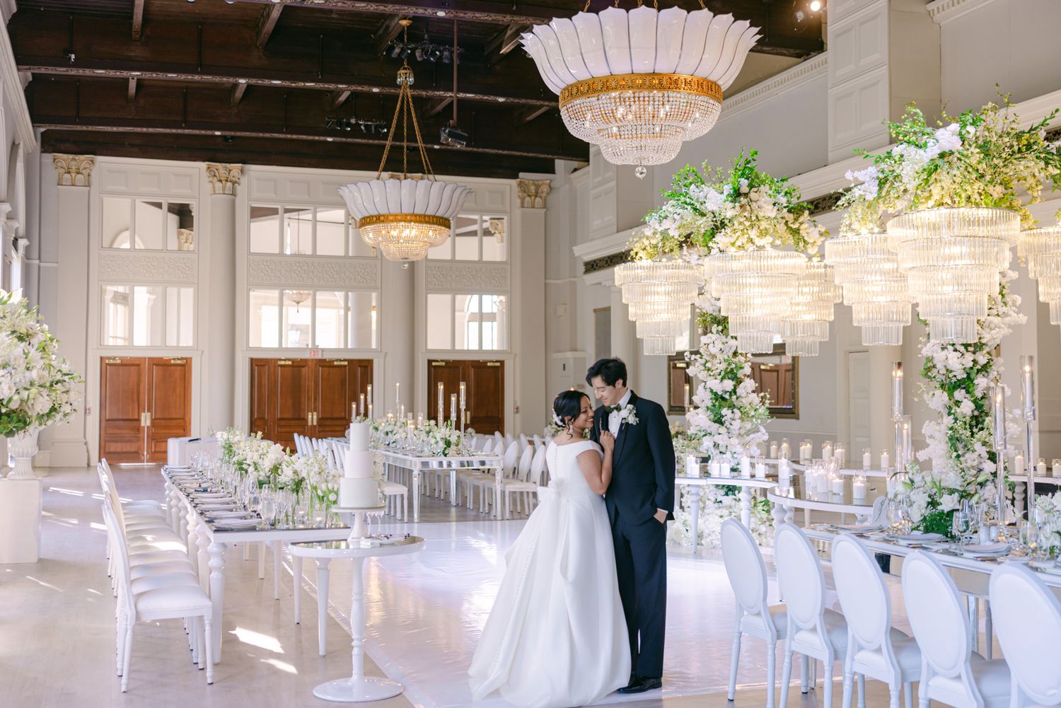 A bride in a white gown and groom in a dark suit embrace in an elegant ballroom with chandeliers, floral centerpieces and white chairs.