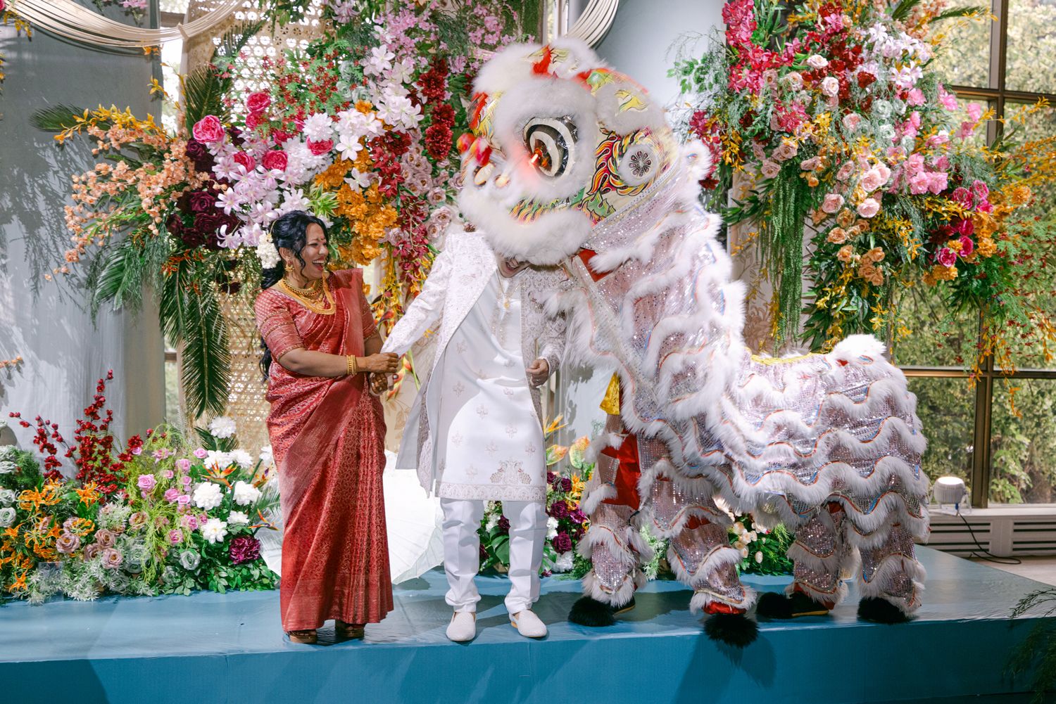 A bride in a red sari stands beside a groom in white as a white lion dance costume performs on a stage with colorful flowers.