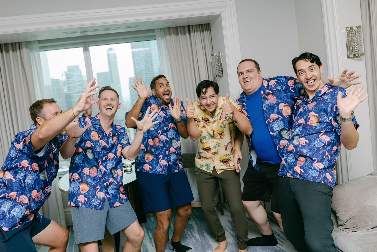 Six cheerful men in matching blue floral Hawaiian shirts cheer and gesture excitedly in a hotel room with a city view.