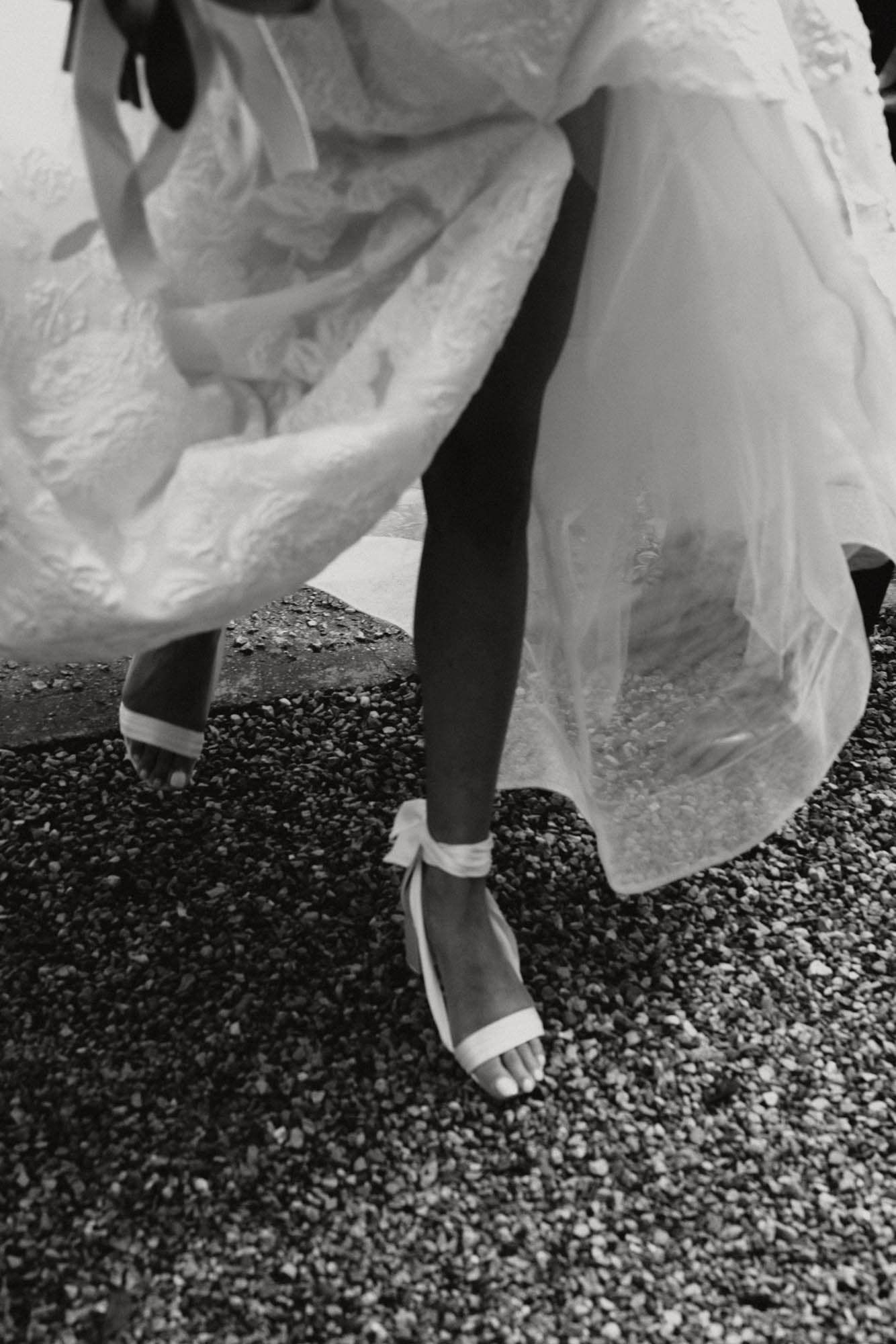 Black and white close-up of a bride adjusting her white strappy heels on a gravel path.