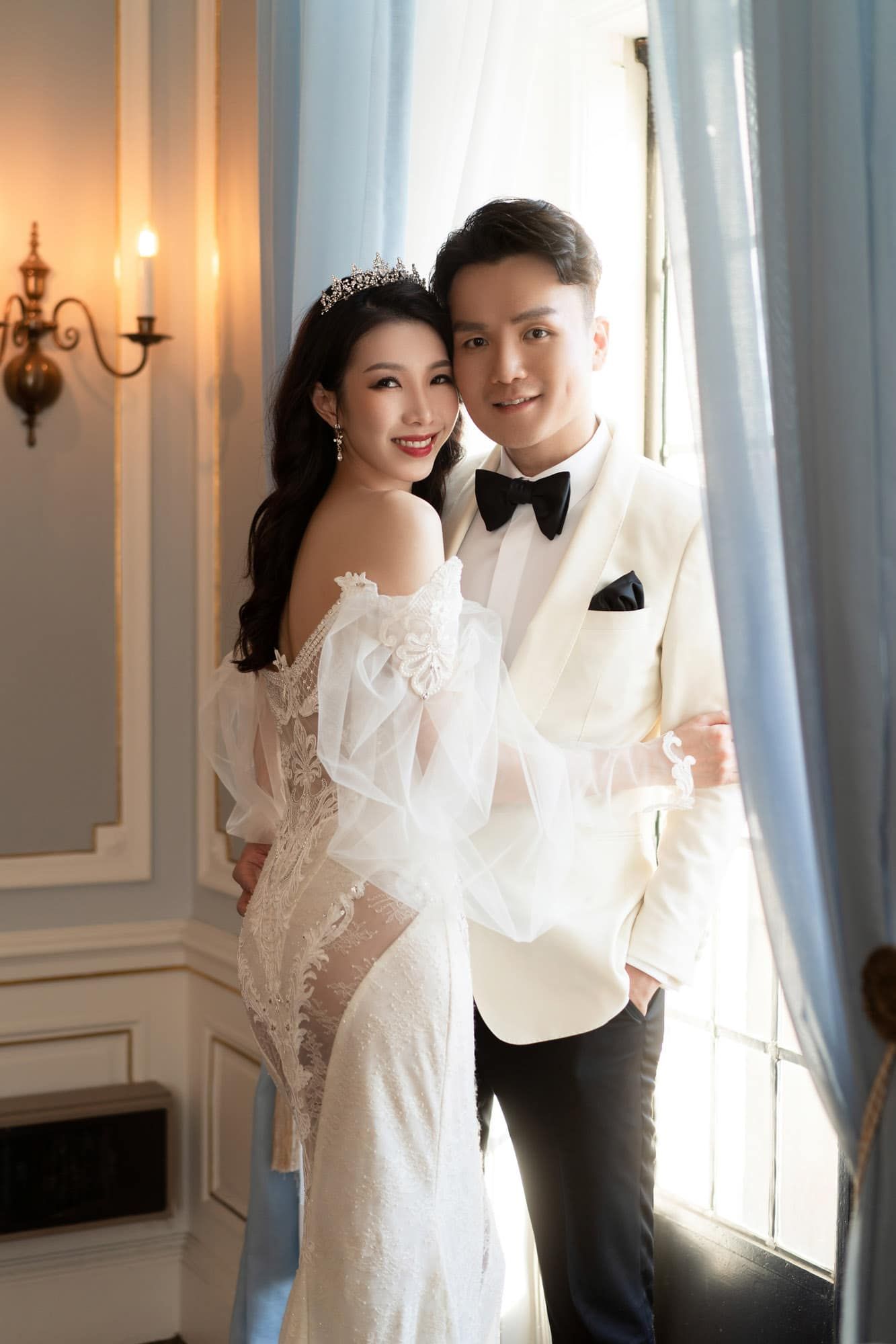 Smiling bride and groom pose together by a draped window in an elegant white-walled room.