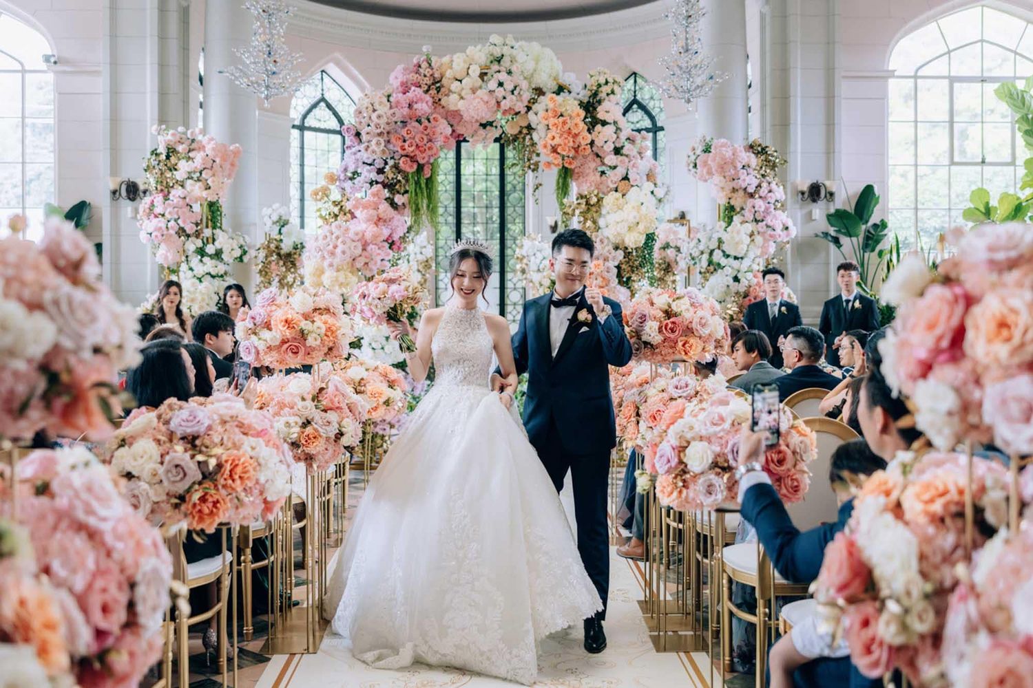 Bride and groom walk joyfully down aisle surrounded by lush pink floral arrangements in chapel.
