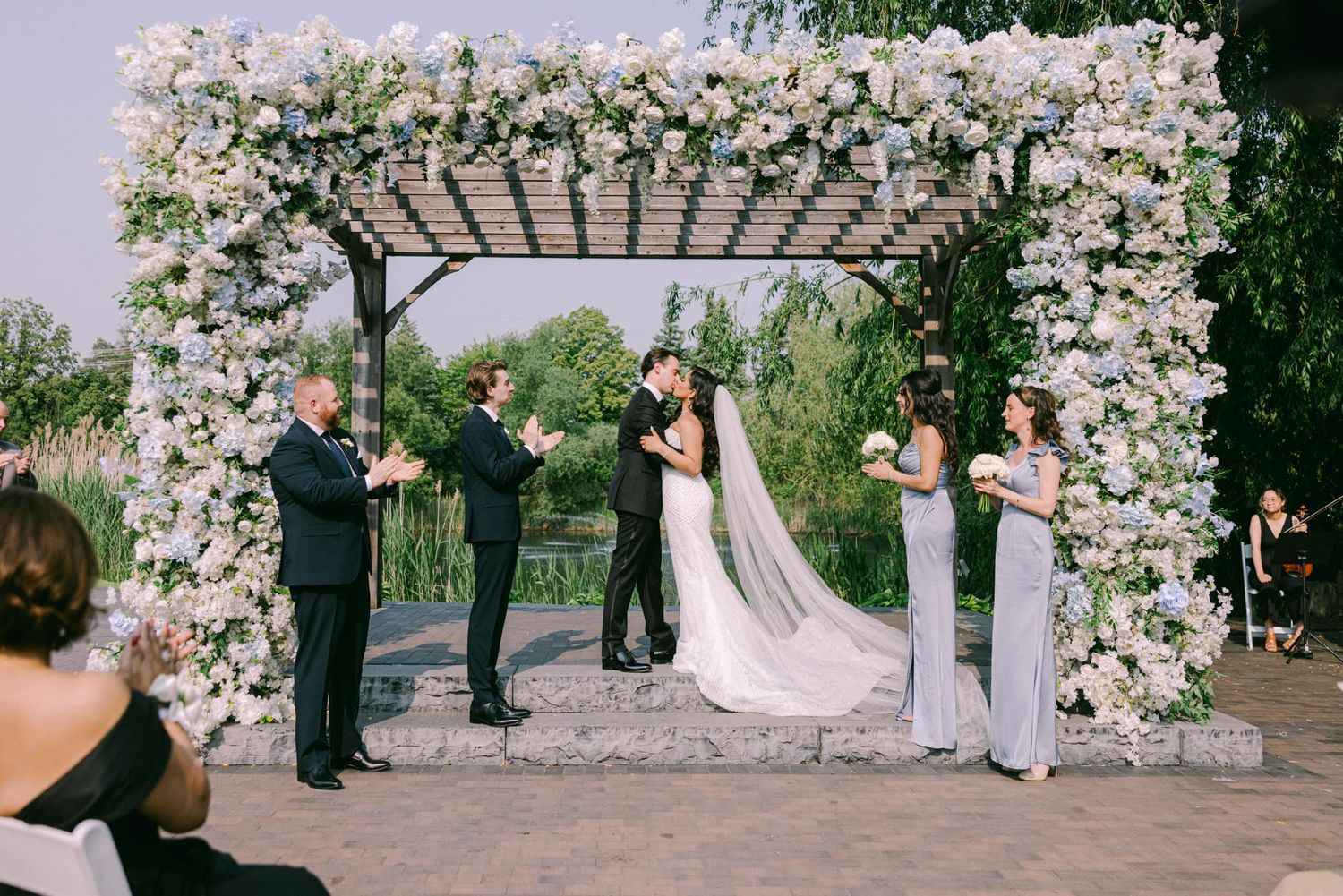 Couple kisses under white floral pergola arch surrounded by wedding guests outdoors.