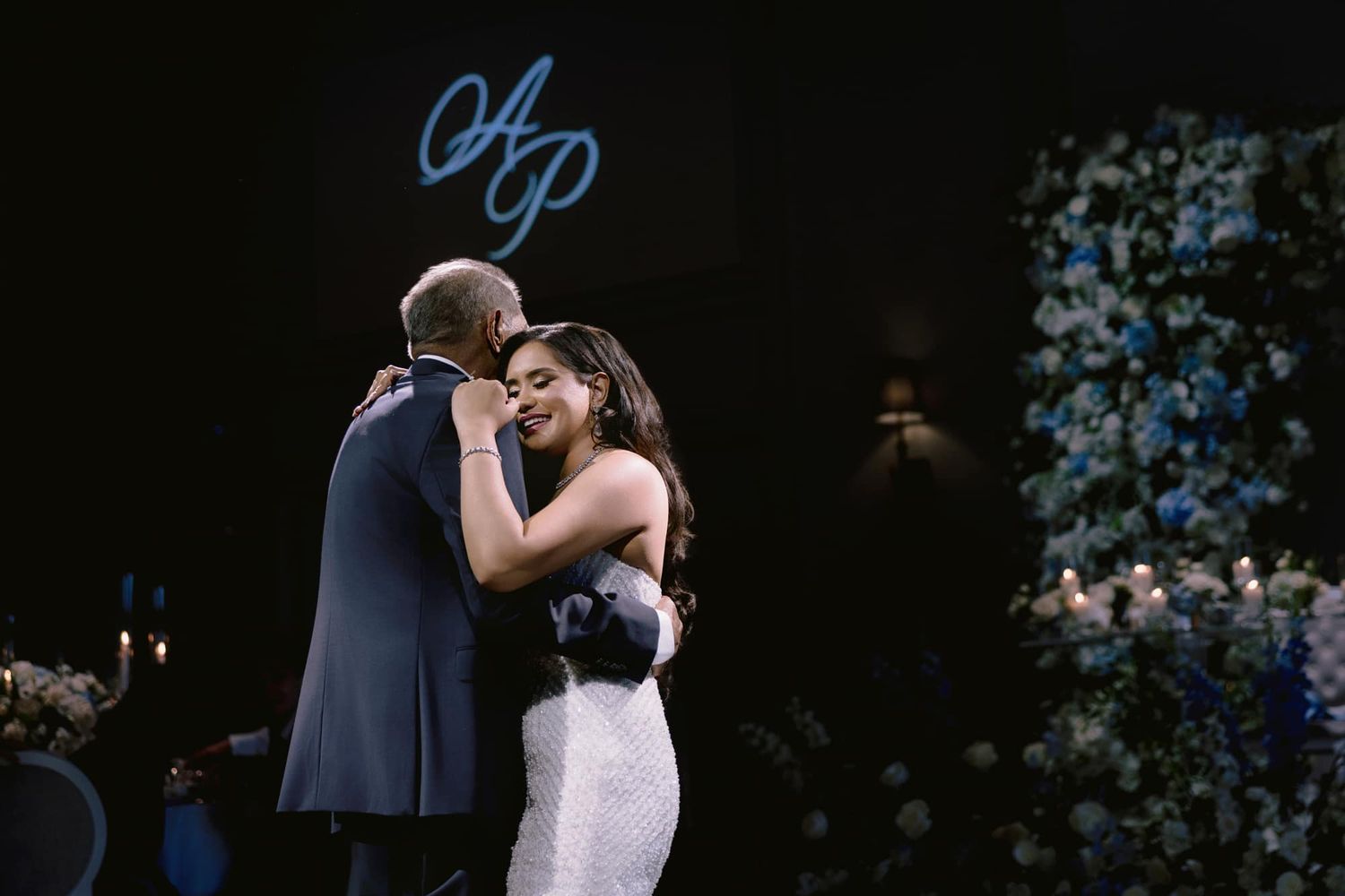 Bride shares emotional father-daughter dance near stunning blue floral wall at wedding reception.