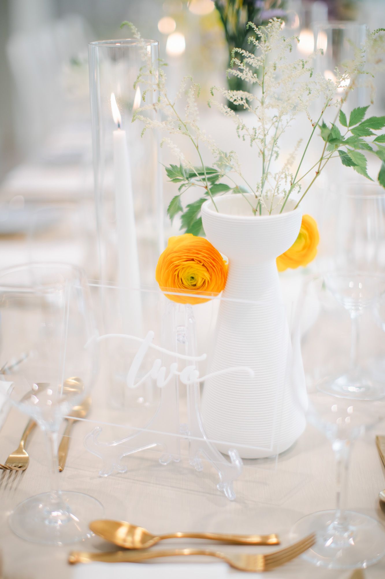 Wedding reception table with orange ranunculus flowers, clear acrylic table number, gold cutlery, and white vase.