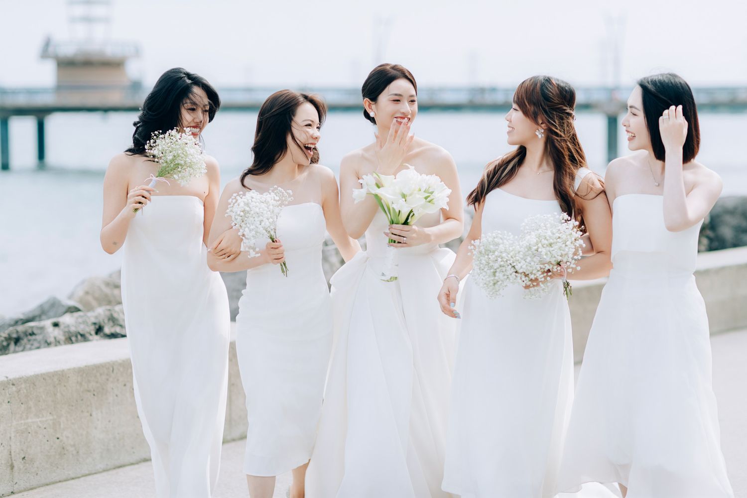 Smiling bride with bridesmaids in white dresses holding baby's breath bouquets by the seaside.