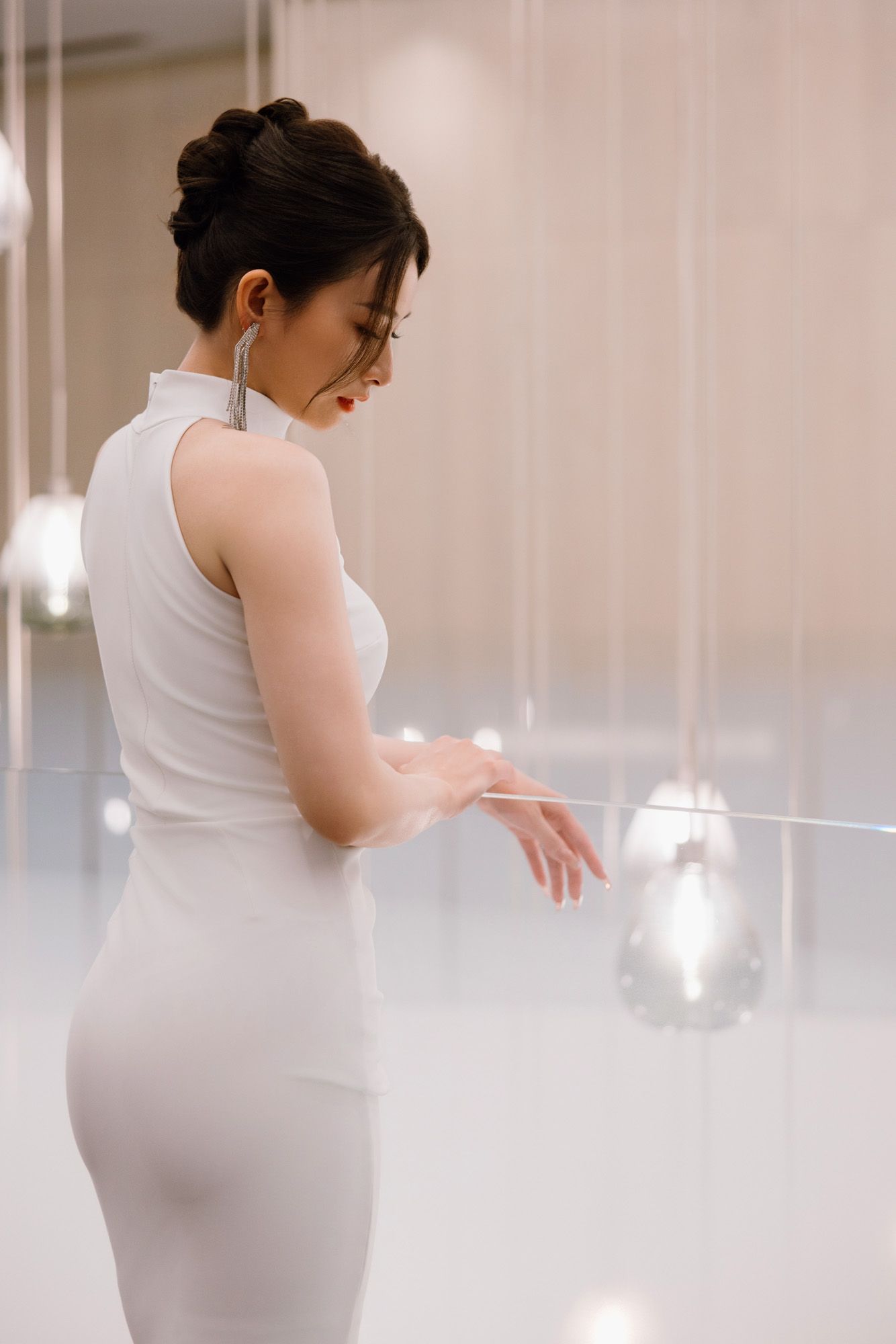 Bride in a fitted white halter gown admires hanging crystal decorations in a softly lit venue.