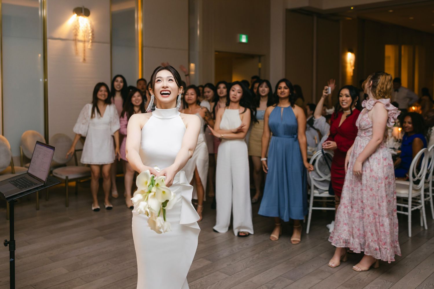Laughing bride in white gown prepares to toss her bouquet to a group of excited female guests.
