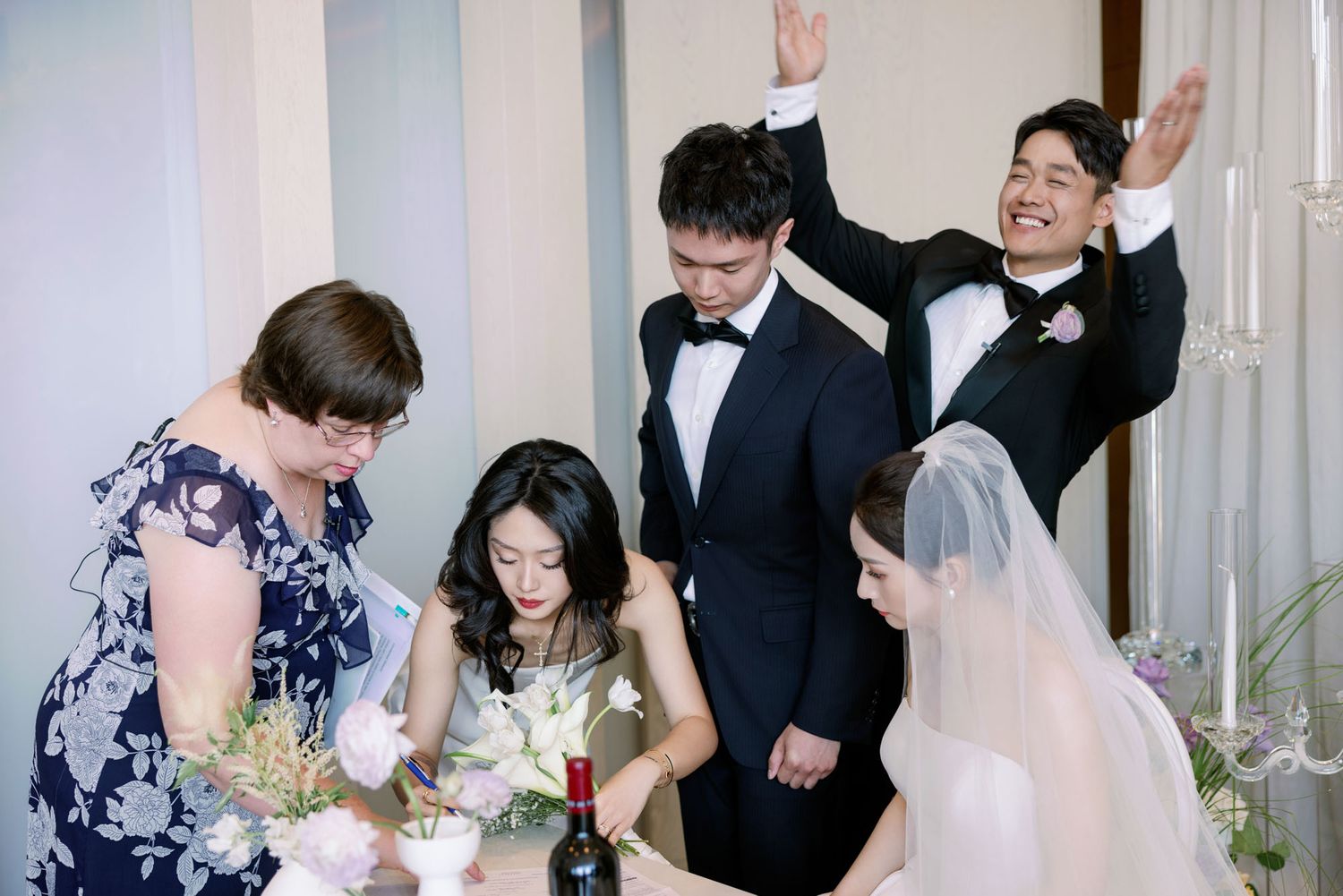 Wedding party gathers around signing table as groom celebrates joyfully at elegant ceremony.