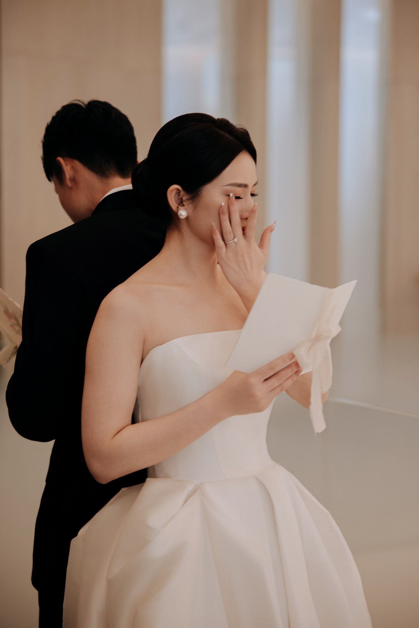 Emotional bride in strapless white gown reads wedding vows letter, covering mouth.