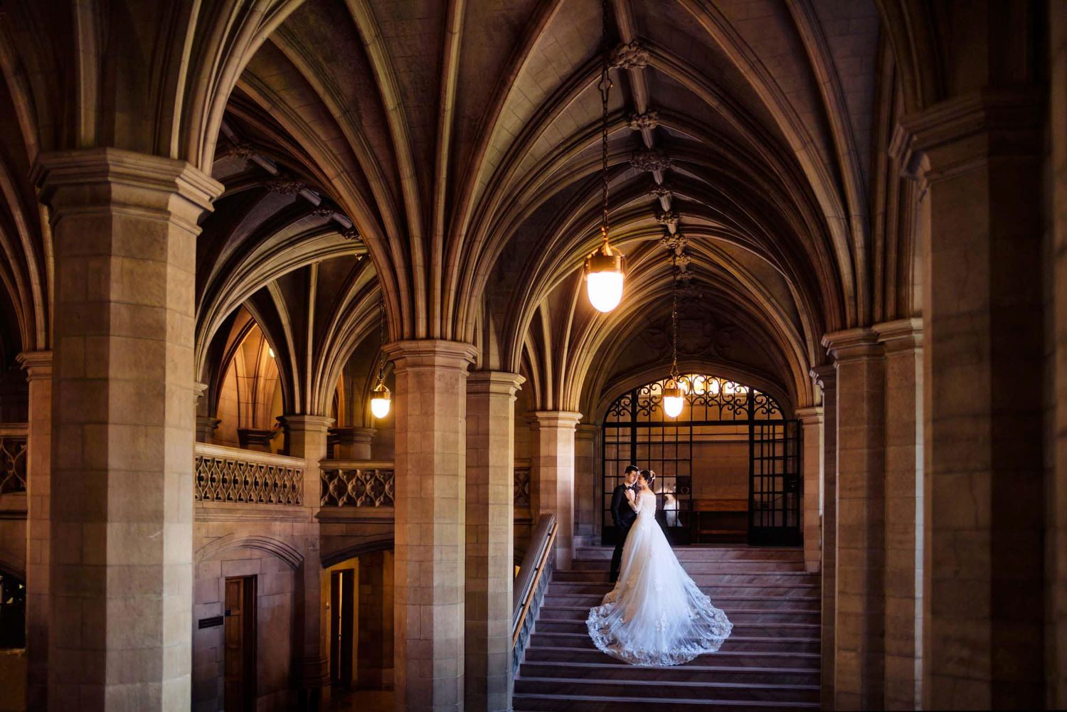 Bride in elegant white gown with long train stands on Gothic arch staircase in historic stone building.