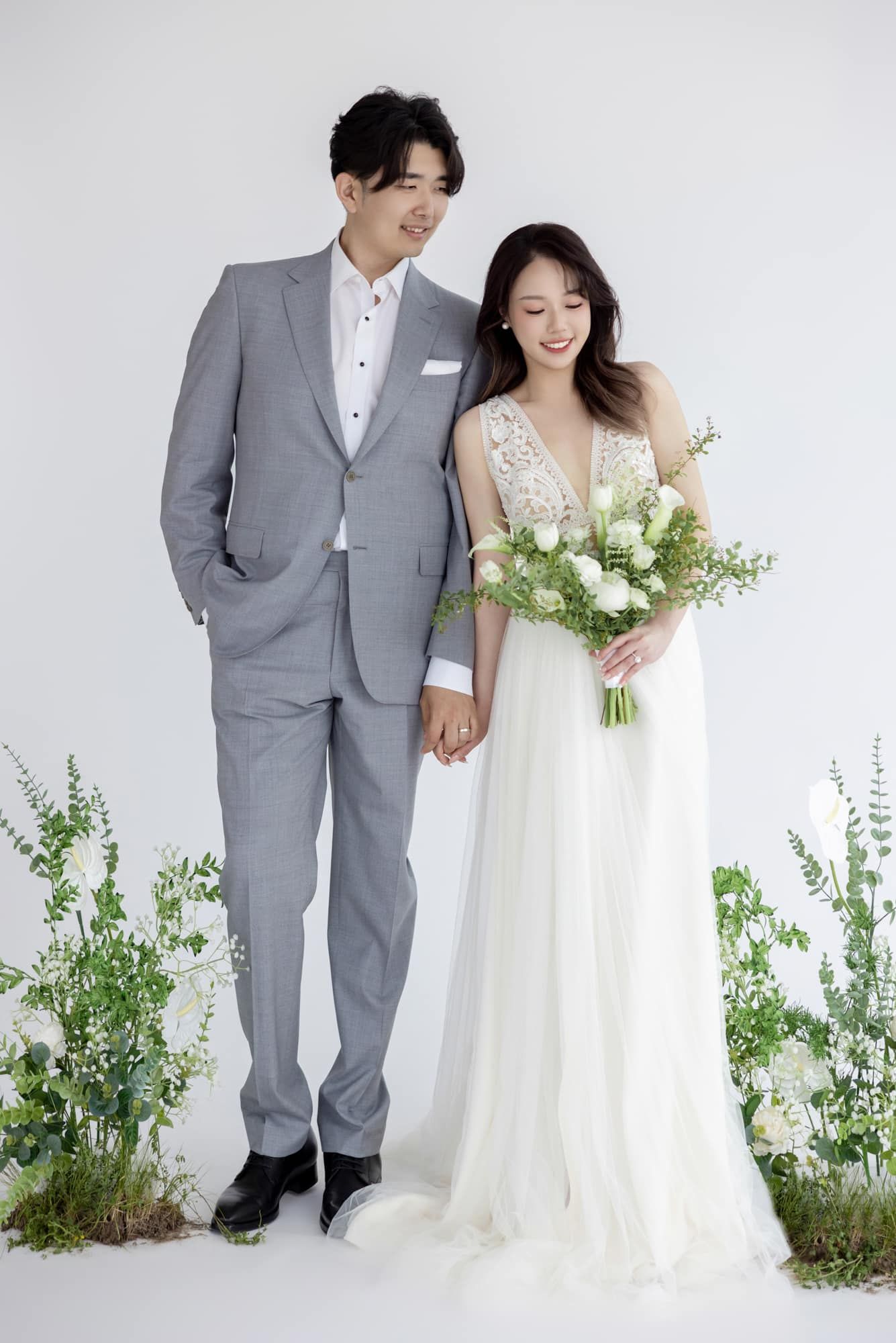 Couple in wedding attire, groom in gray suit, bride in white gown holding bouquet, posing with greenery backdrop.