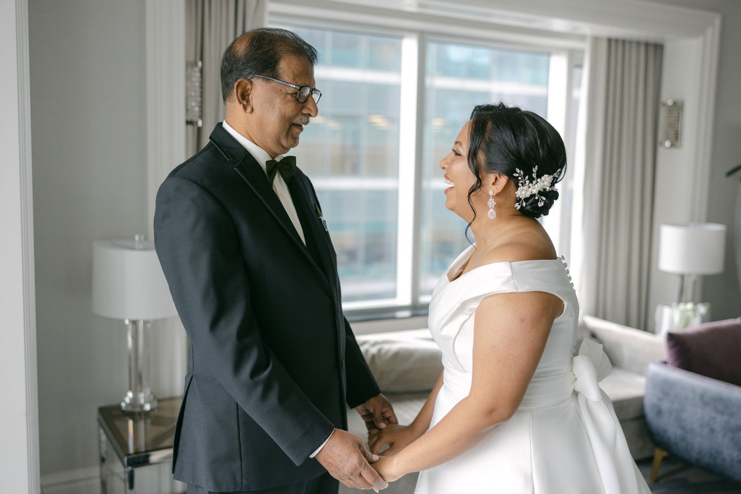 A bride in an off-the-shoulder white gown holds hands with a smiling man in a dark suit beside a bright hotel room window.