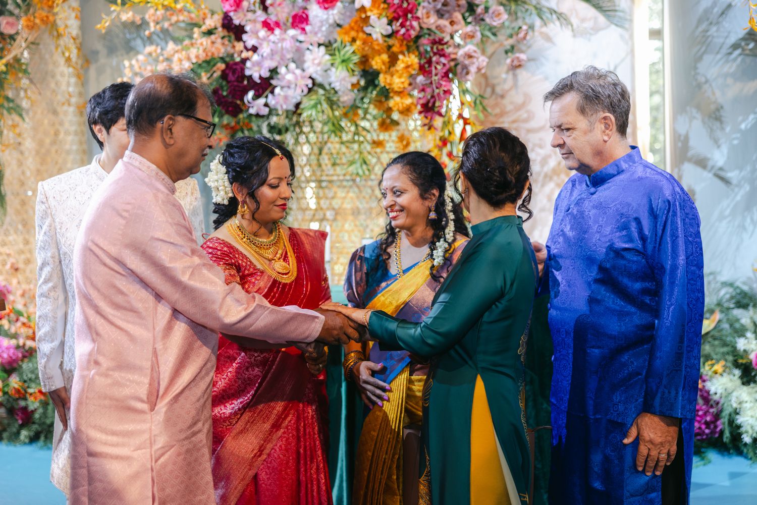 Two families join hands in an Indian wedding ceremony in colorful saris and festive attire before a backdrop of orange and pink flowers.