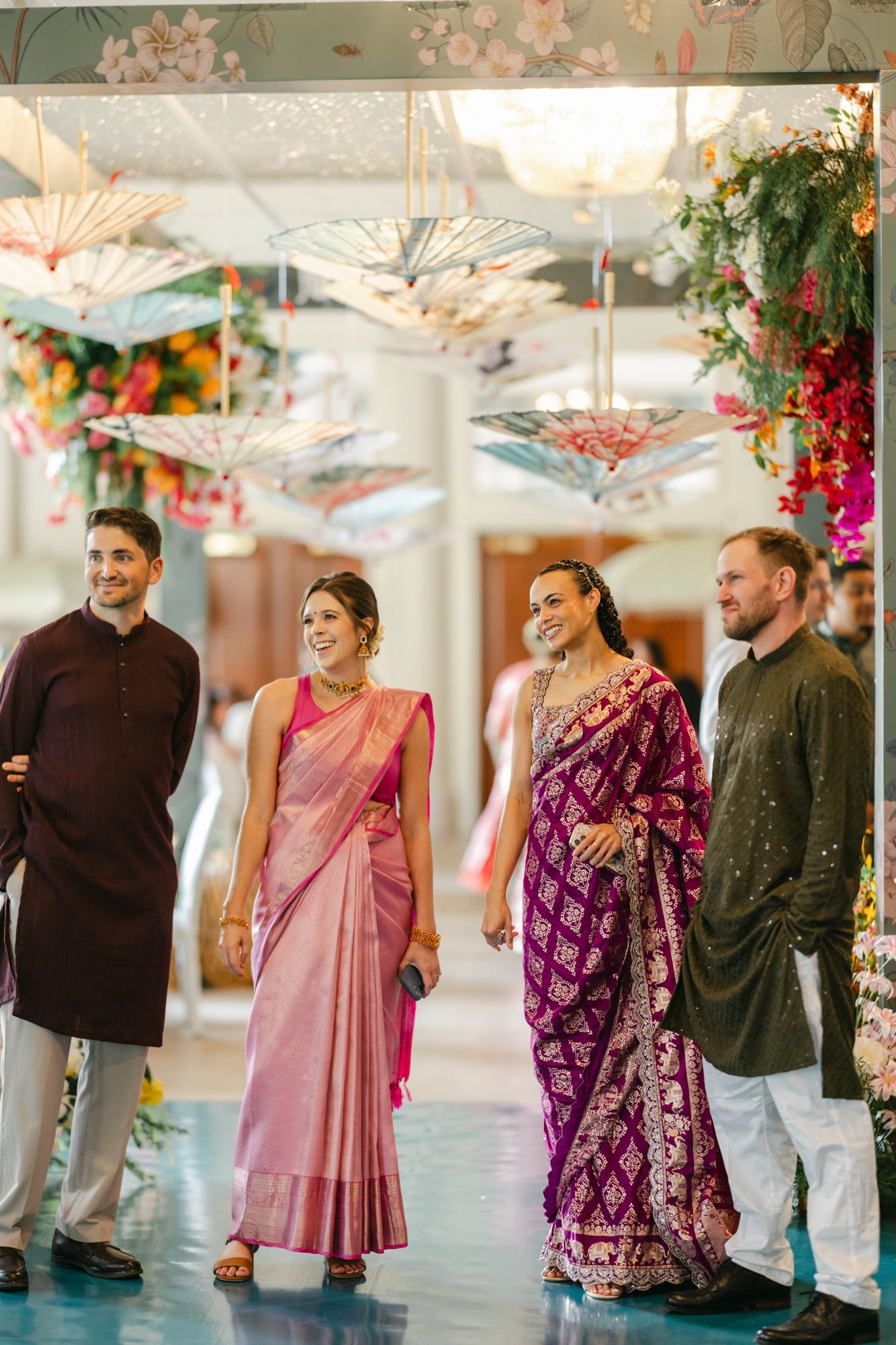 Two women in pink and purple silk sarees laugh beside two men in kurtas at a wedding venue with hanging umbrellas and floral decor.